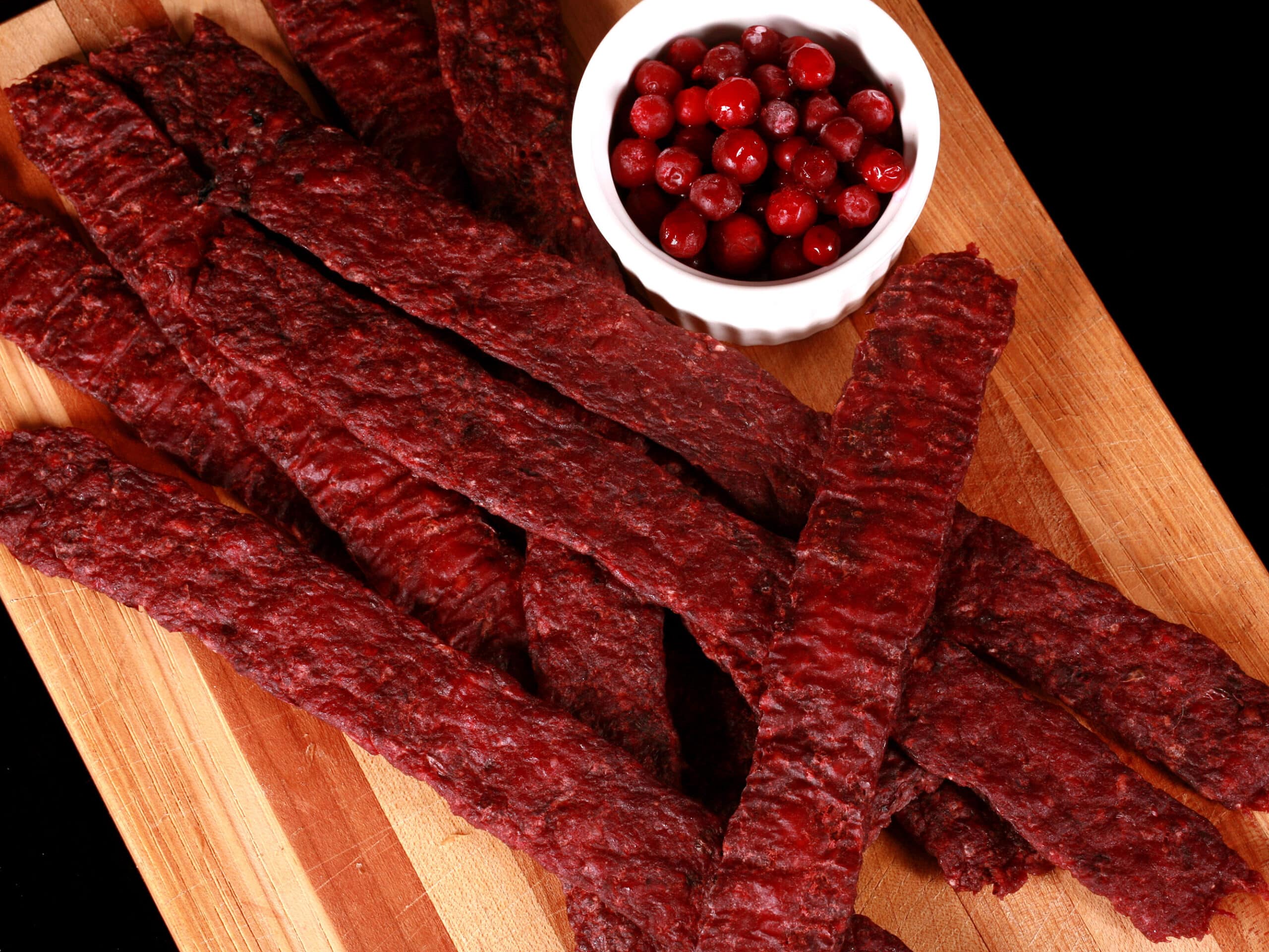 Several strips of homemade lingonberry elk jerky on a wooden cutting board, along with a small bowl of partridgeberries.