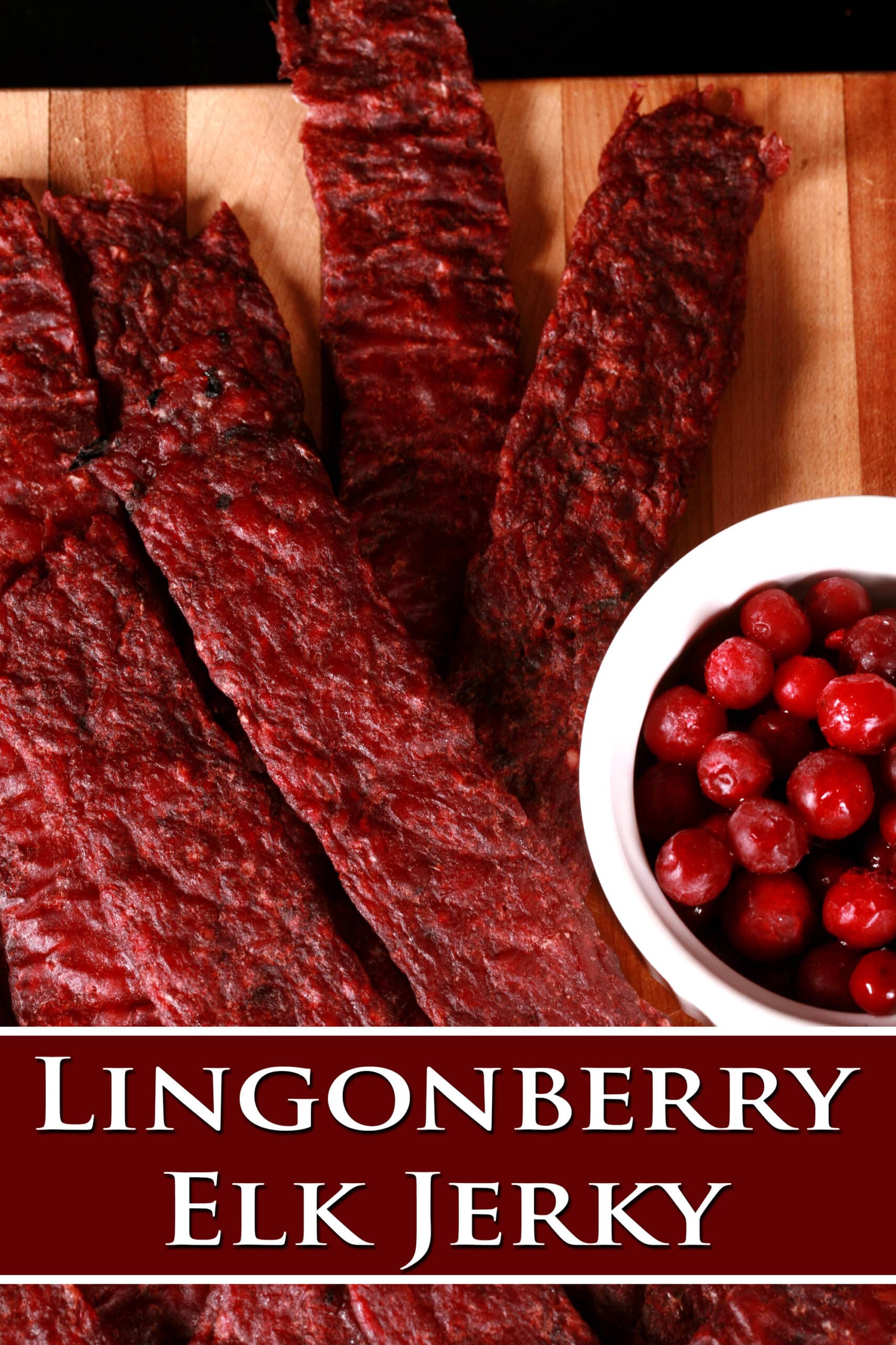 Several strips of homemade elk jerky on a wooden cutting board, along with a small bowl of lingonberries. Overlaid text says lingonberry elk jerky.