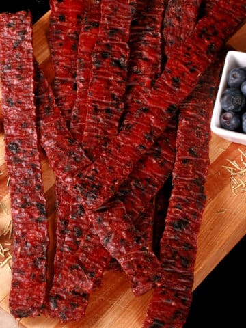 A pile of homemade rosemary blueberry ground beef jerky on a wooden cutting board with a bowl of fresh blueberries.