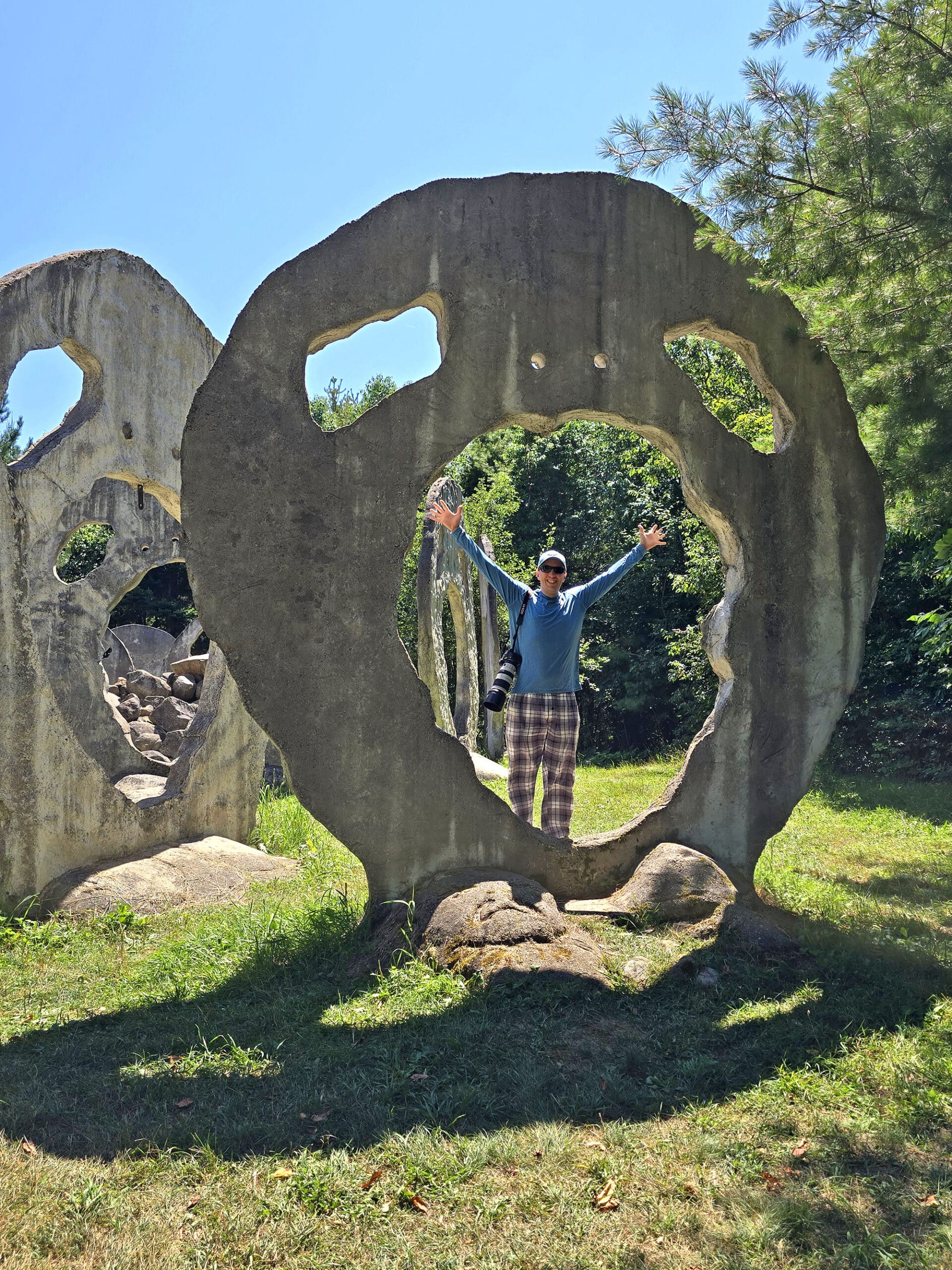 A grassy area with several 18 foot tall concrete sculptures that look like screaming heads. The author’s husband is standing in front of one of them.