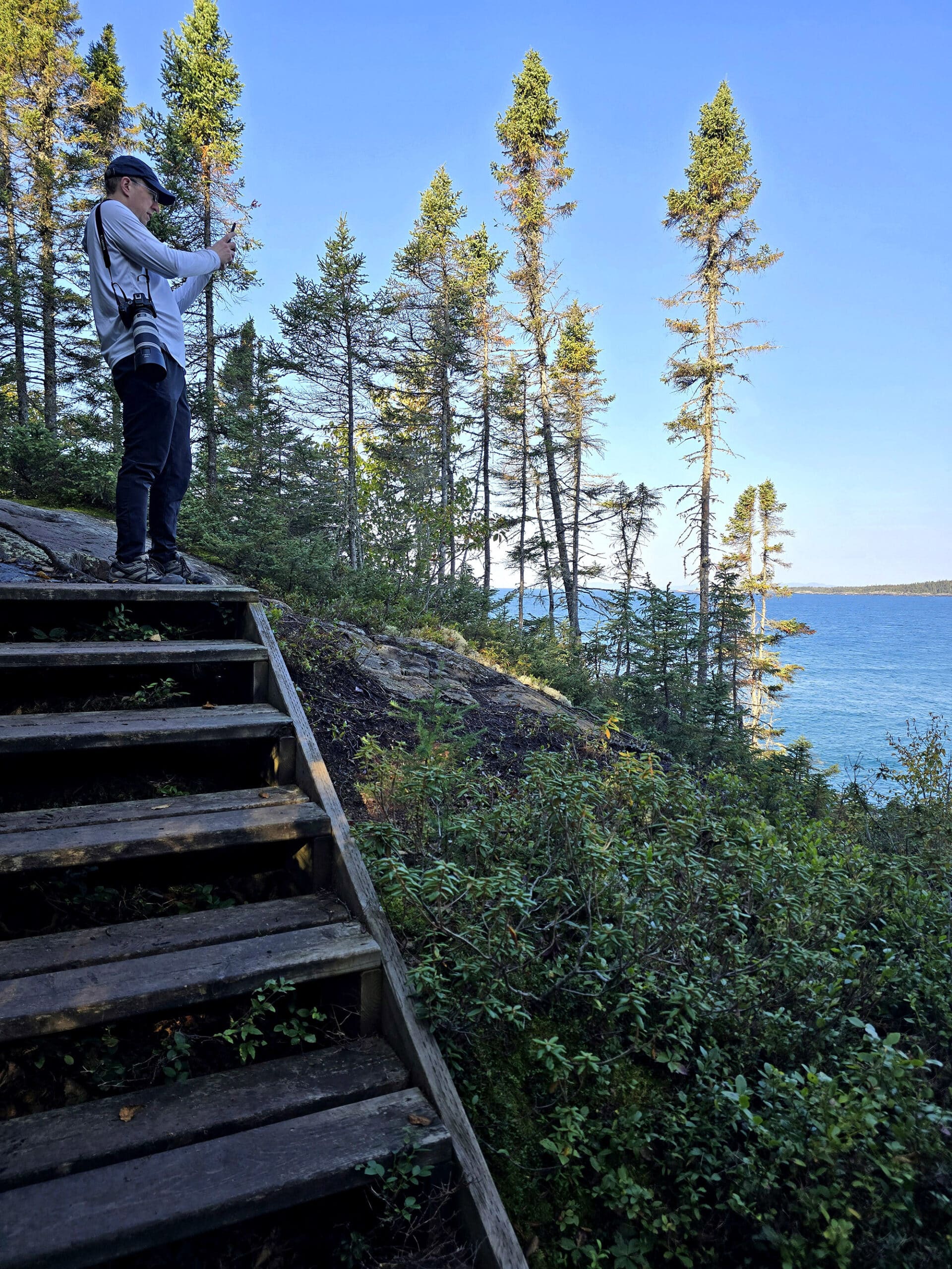 A man photographing lake superior from atop a set of wooden steps on a cliff.