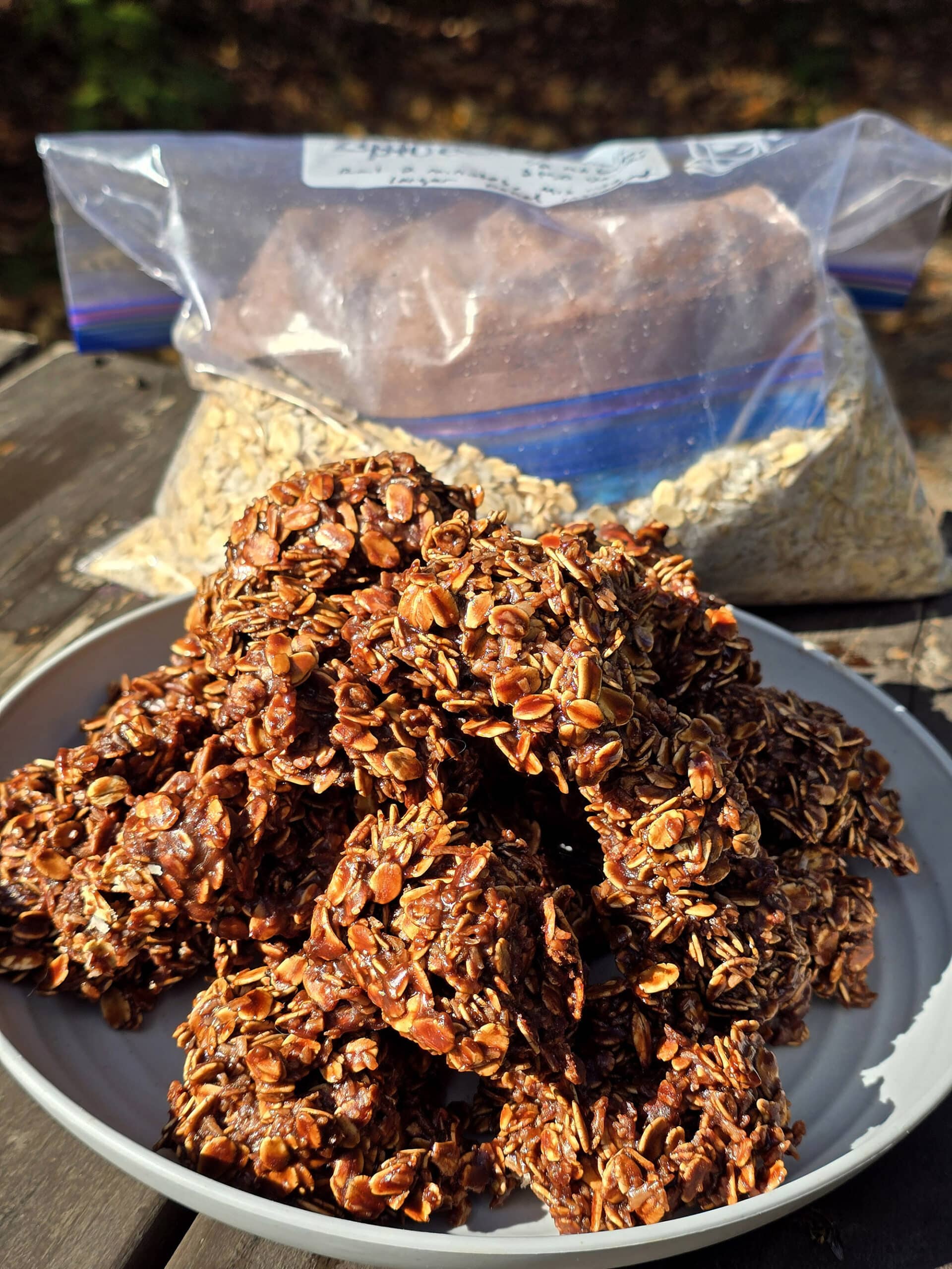 A plate of campfire chocolate haystack cookies on a picnic table, in front of a bag of homemade cookie mix.