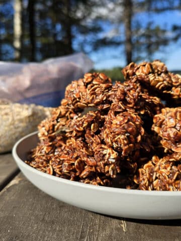 A plate of campfire chocolate haystack cookies on a picnic table, in front of a bag of homemade cookie mix.