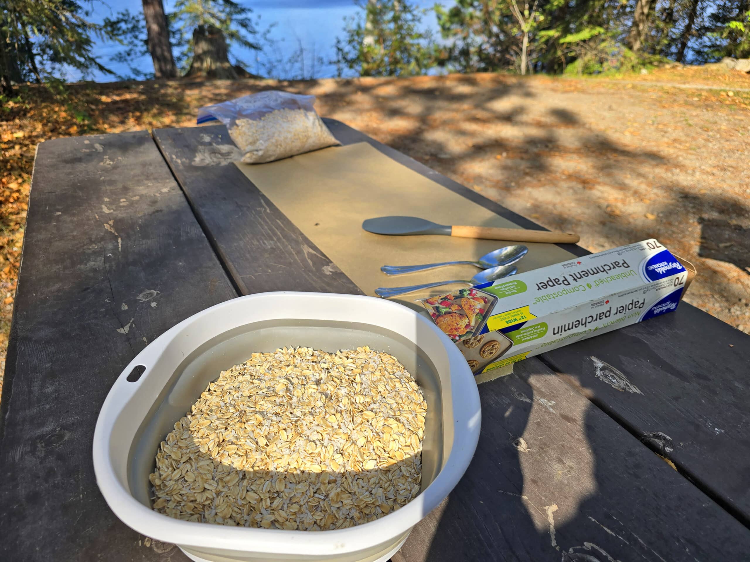 A bowl of oat mixture and parchment paper spread out on a picnic table in front of a lake.