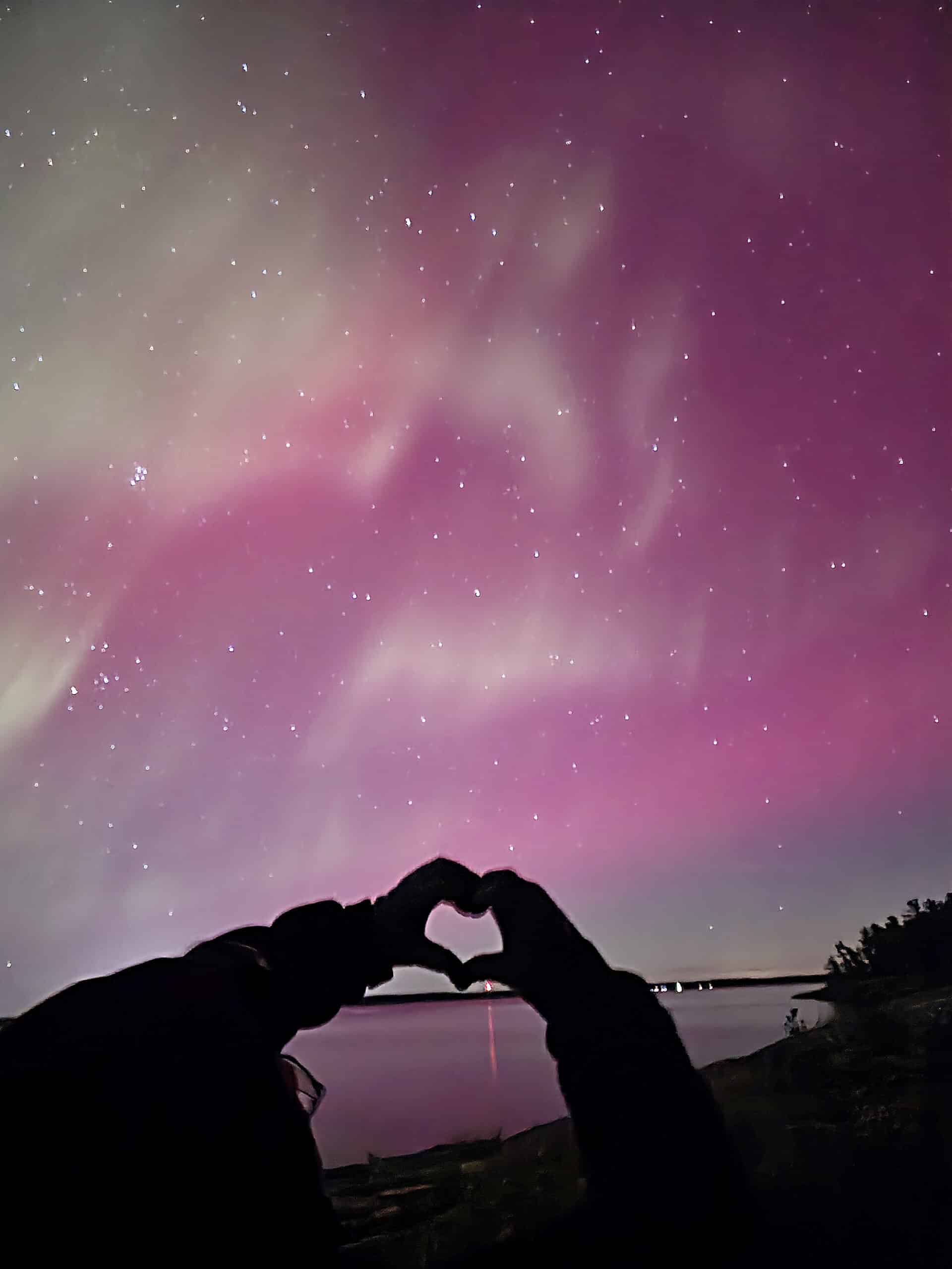 A pair of hands forming a heart in front of pink northern lights over a lake.