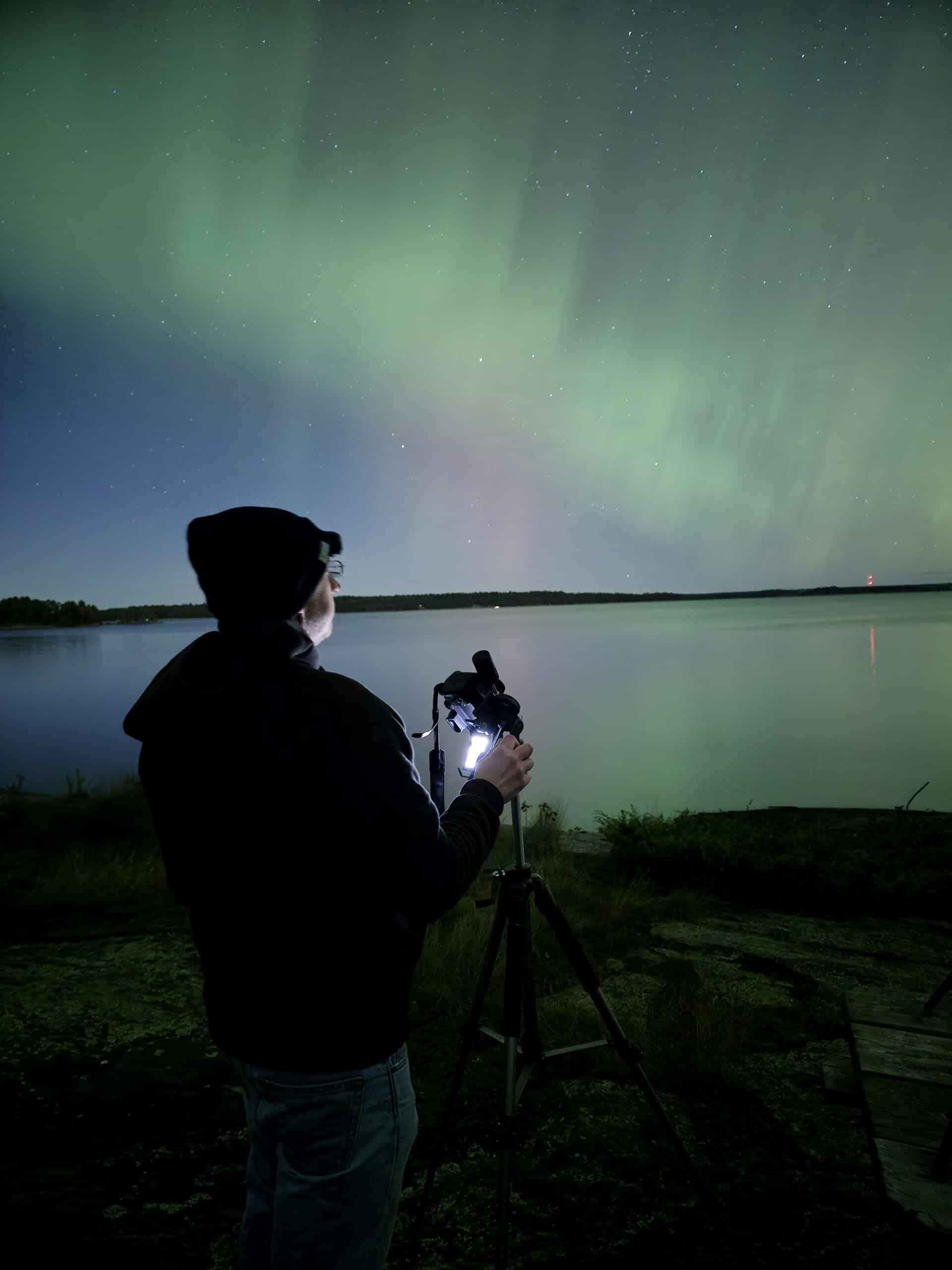 A man and a camera in front of a lake, with northern lights illuminating the sky.