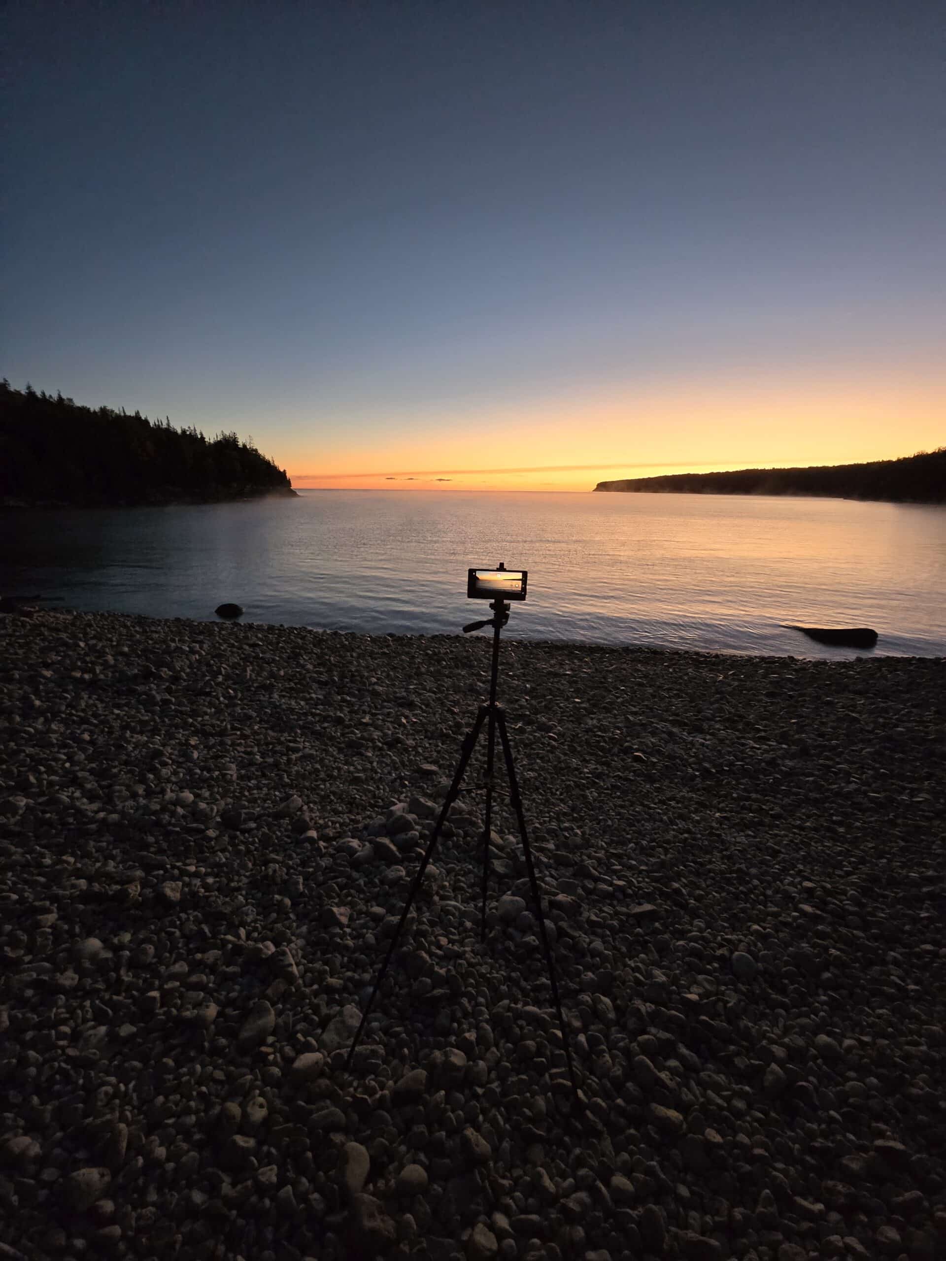 A beach scene at dawn. A cell phone on a tripod is capturing a photo of the scene.