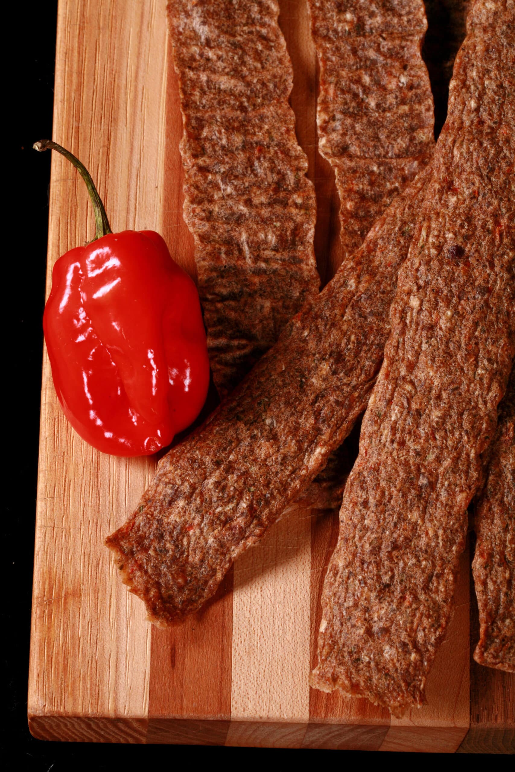 Several strips of jerk chicken jerky on a cutting board, with green onions and scotch bonnet peppers.