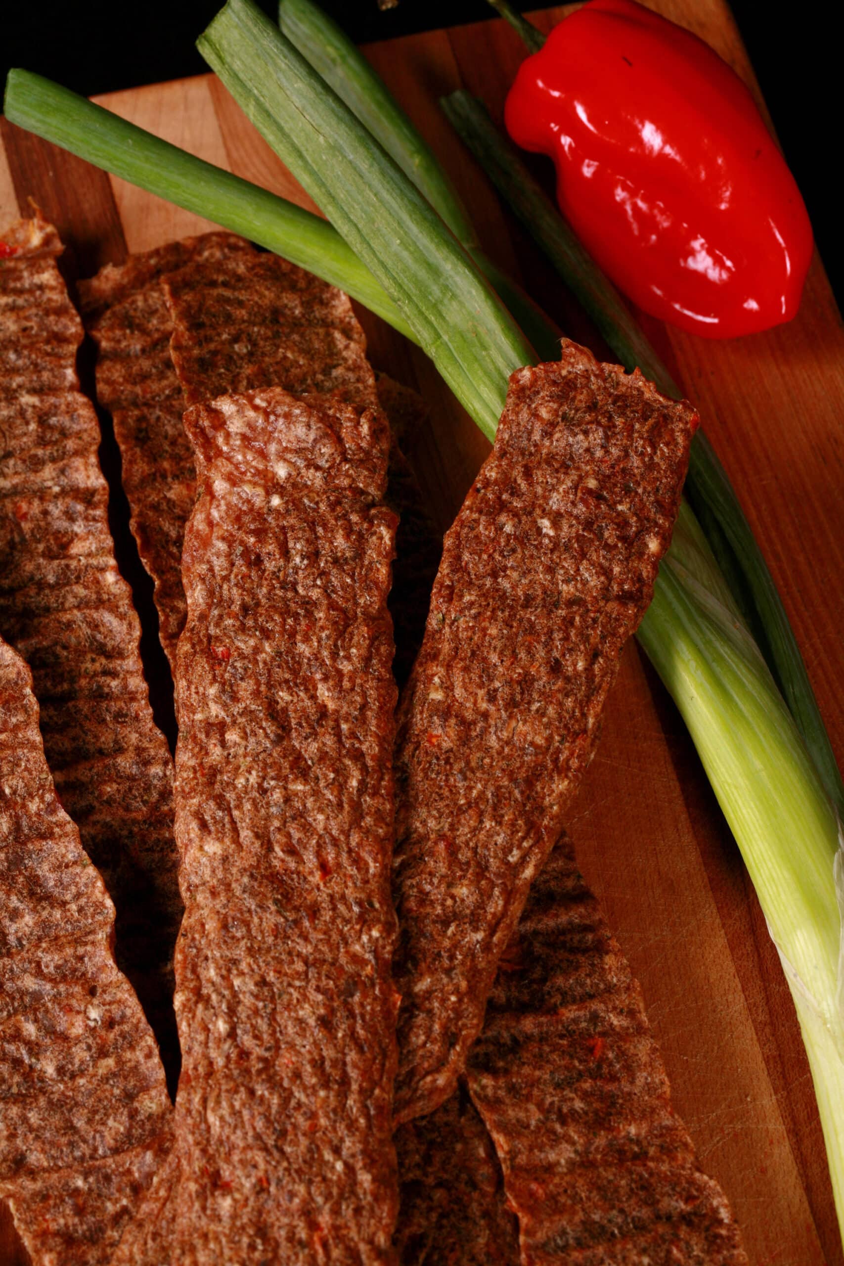 Several strips of Jamaican jerk chicken jerky on a cutting board, with green onions and scotch bonnet peppers.