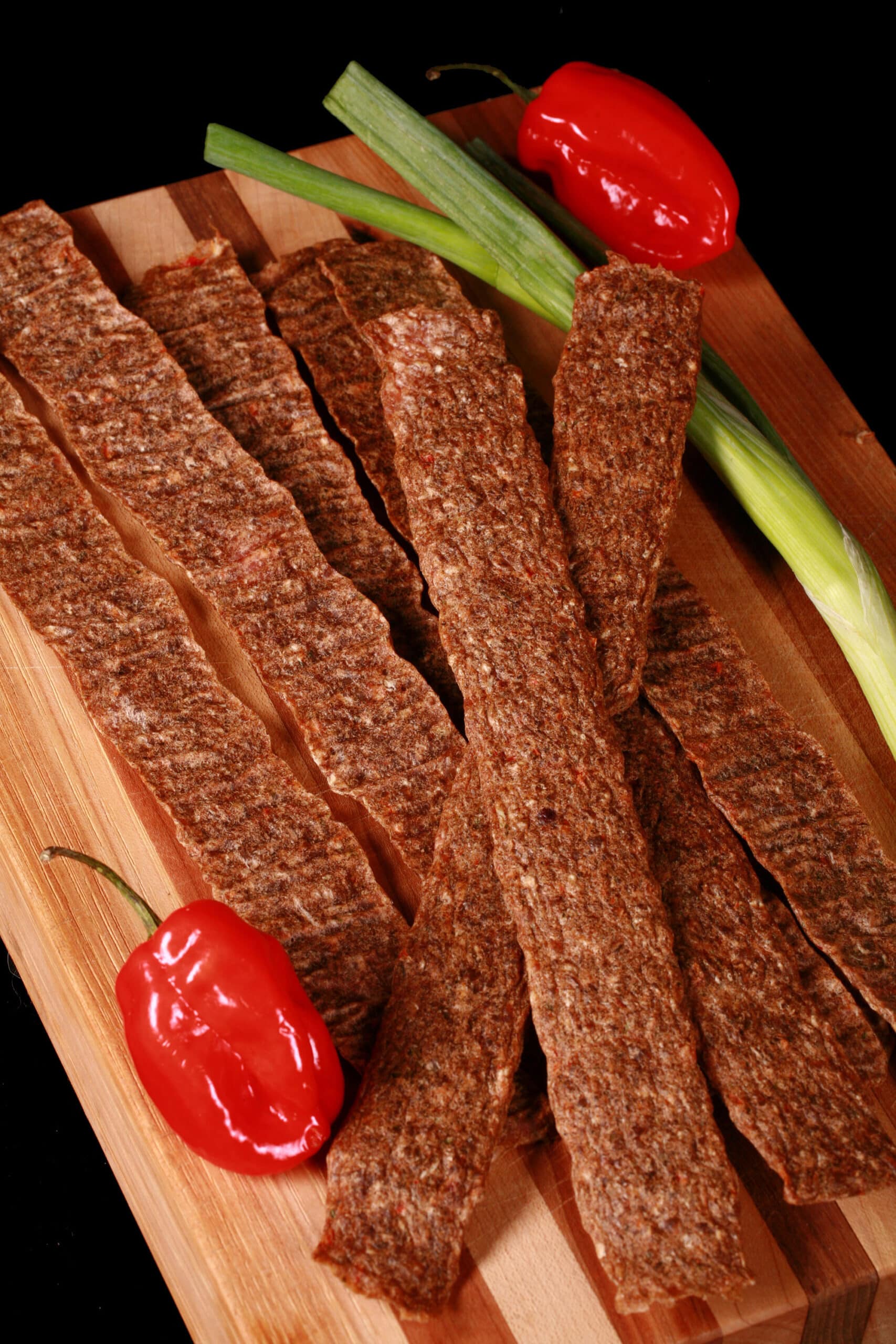 Several strips of jerk chicken jerky on a cutting board, with green onions and scotch bonnet peppers.