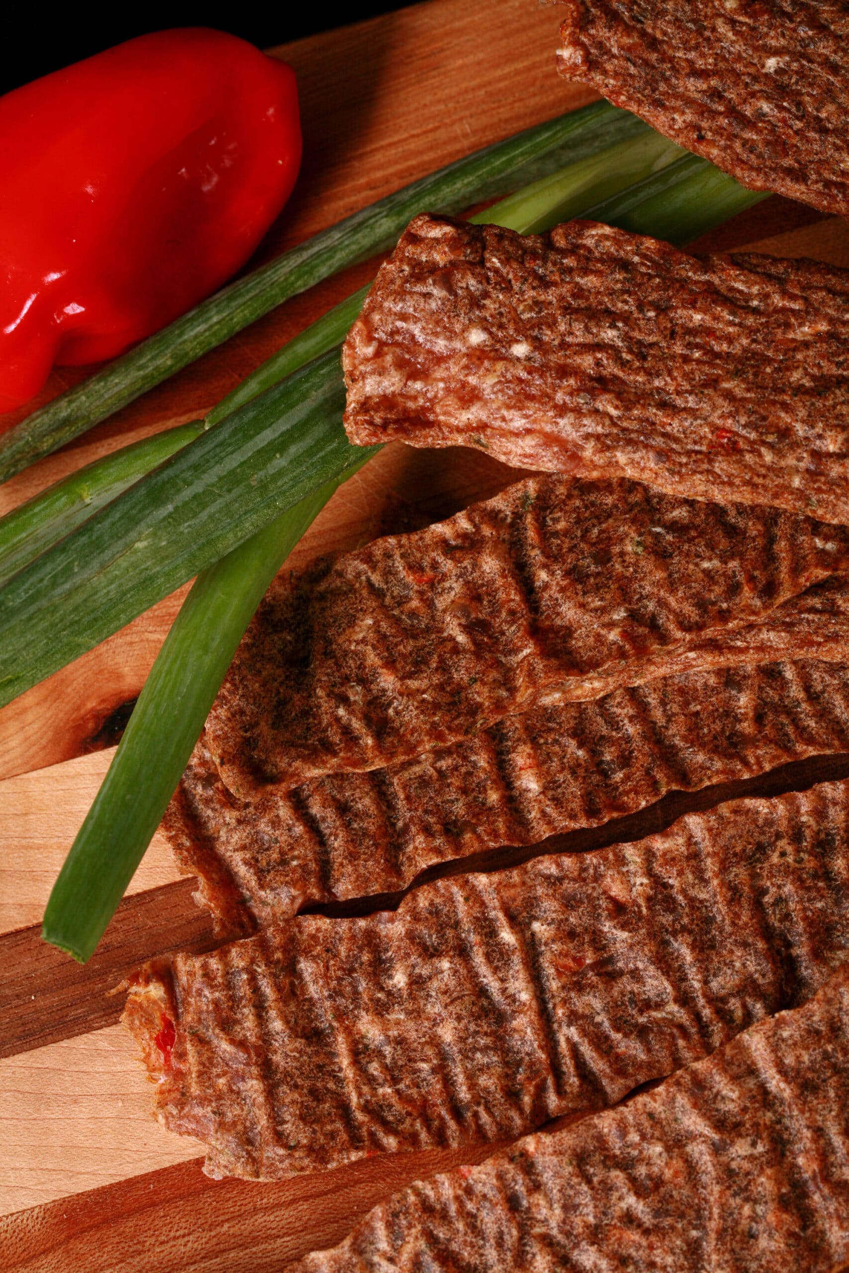 Several strips of jerk chicken jerky on a cutting board, with green onions and scotch bonnet peppers.