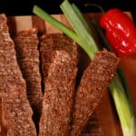 Several strips of Jamaican jerk chicken jerky on a cutting board, with green onions and scotch bonnet peppers.