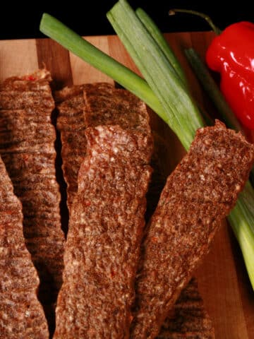 Several strips of Jamaican jerk chicken jerky on a cutting board, with green onions and scotch bonnet peppers.