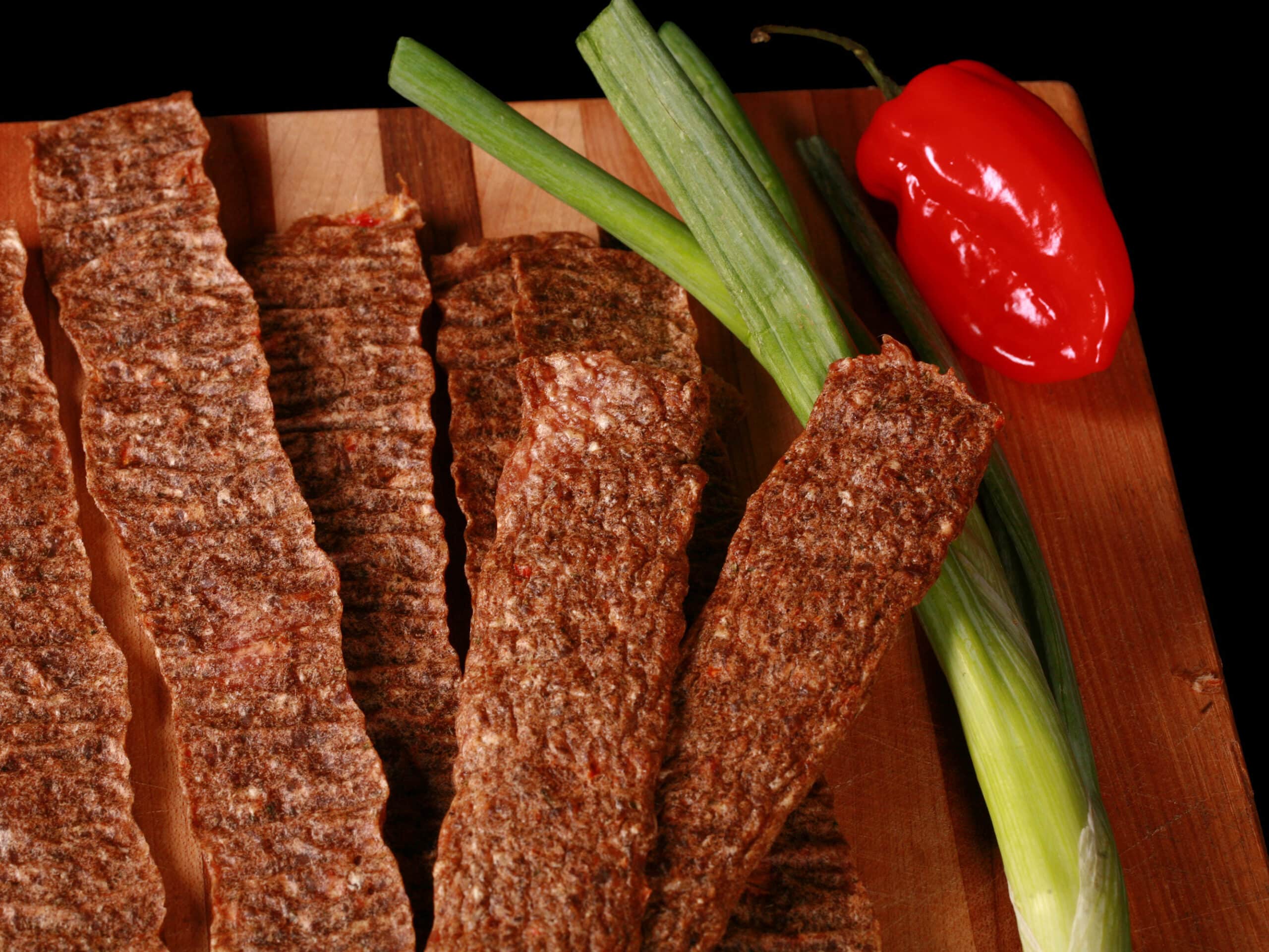 Several strips of Jamaican jerk chicken jerky on a cutting board, with green onions and scotch bonnet peppers.