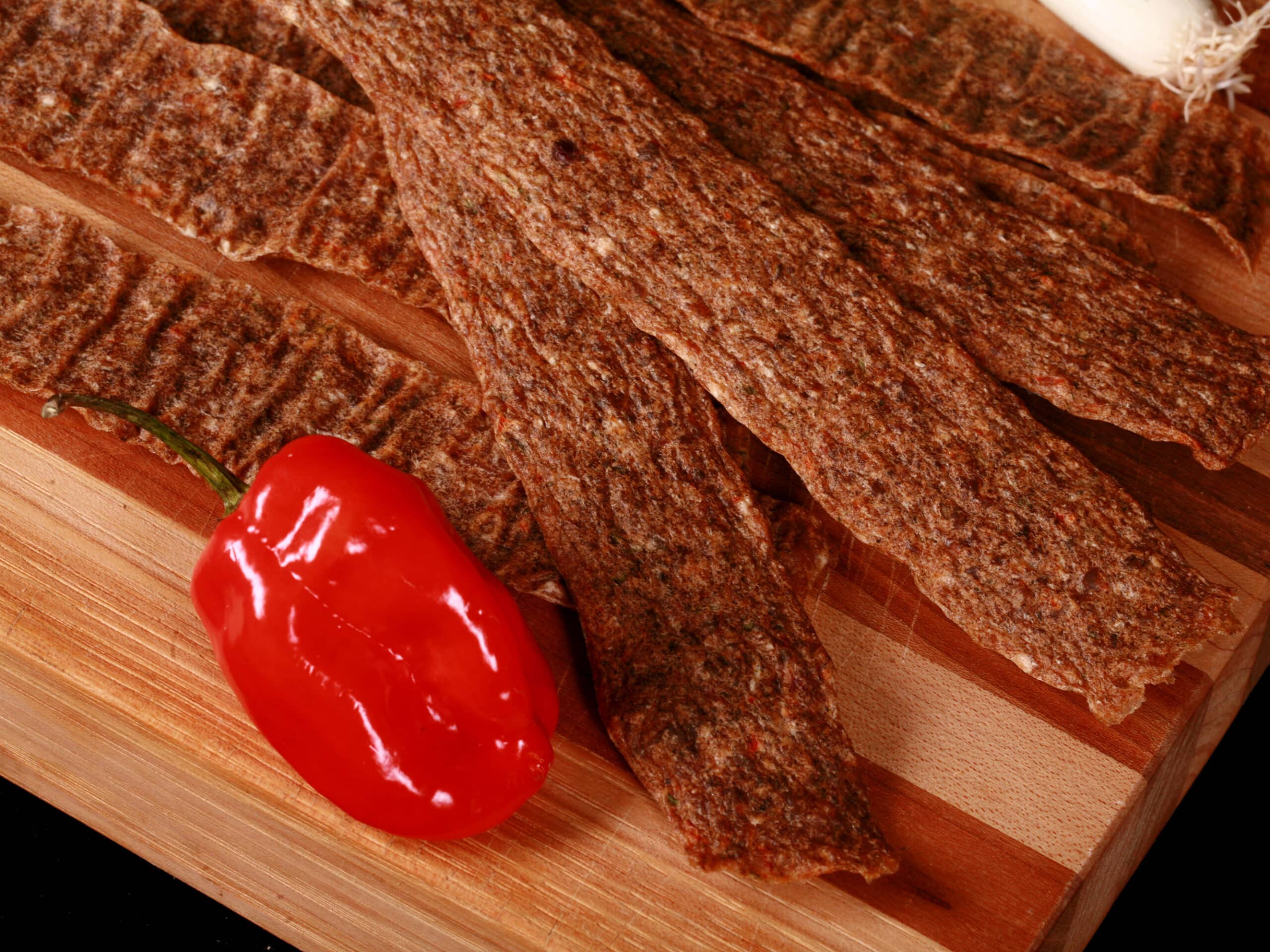 Several strips of jerk chicken jerky on a cutting board, with green onions and scotch bonnet peppers.