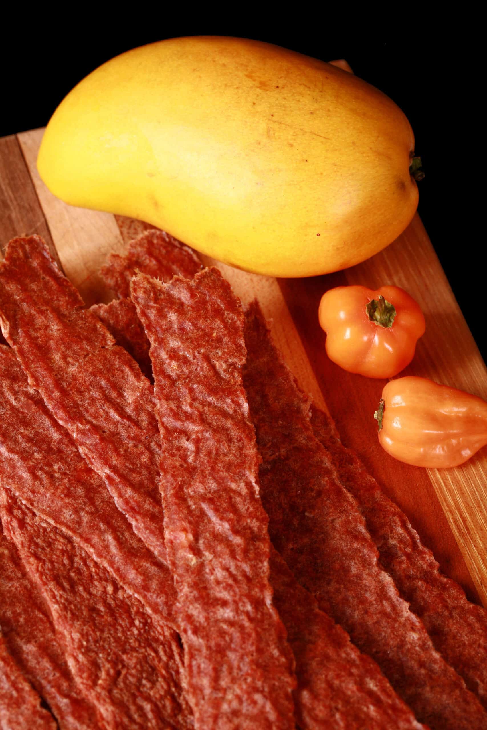 A pile of mango habanero chicken jerky on a cutting board with a mango and 2 habanero peppers.