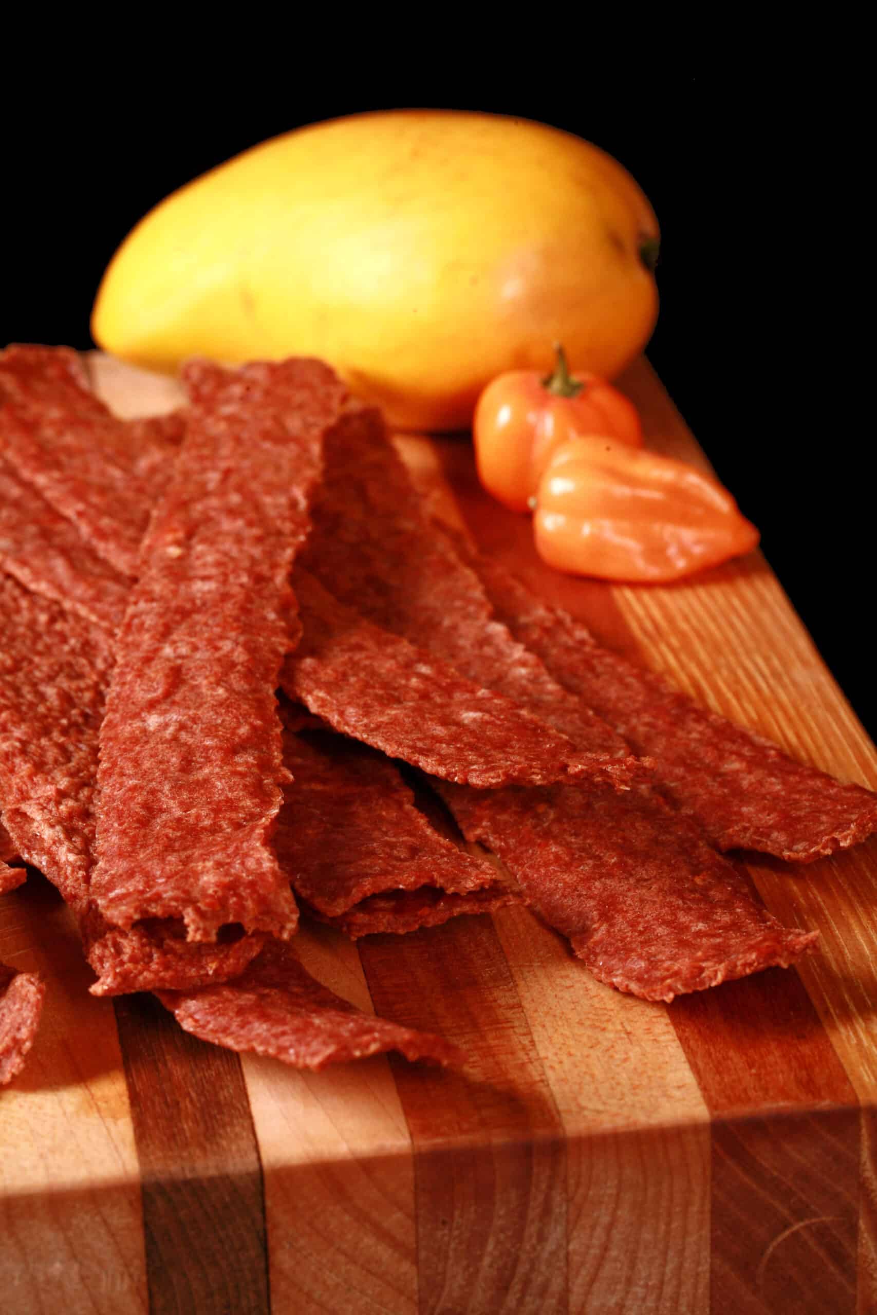 A pile of mango habanero chicken jerky on a cutting board with a mango and 2 habanero peppers.