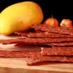 A pile of mango habanero chicken jerky on a cutting board with a mango and 2 habanero peppers.