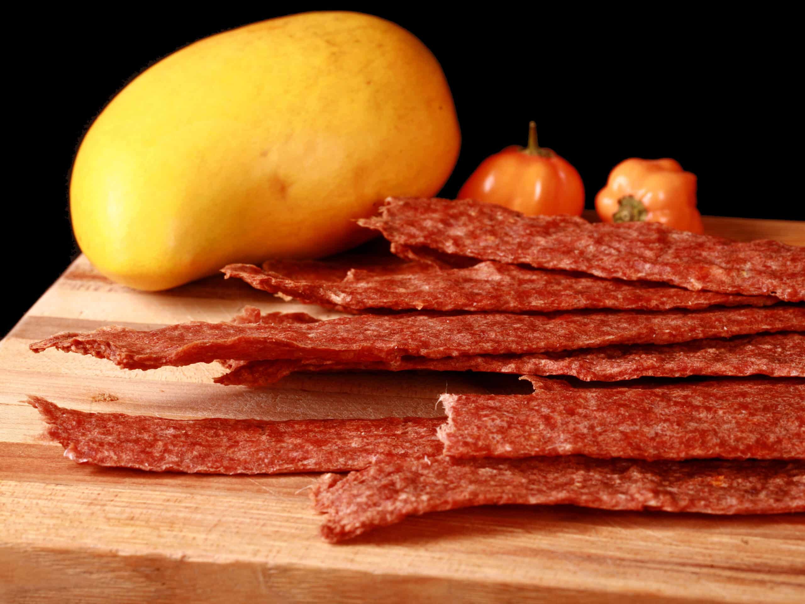 A pile of mango habanero chicken jerky on a cutting board with a mango and 2 habanero peppers.