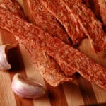 Several strips of homemade chicken jerky on a cutting board with some garlic.