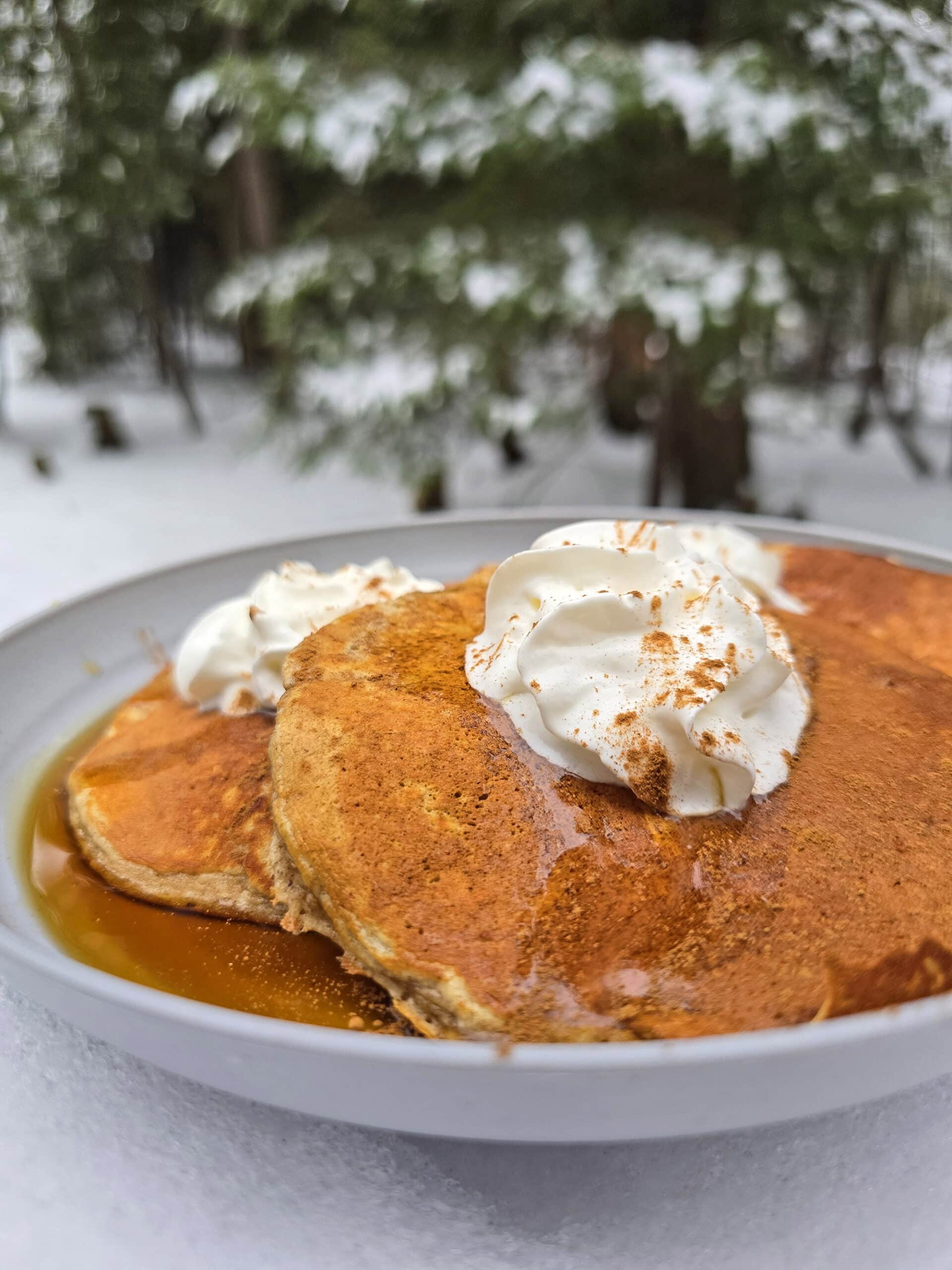 A plate of gingerbread protein pancakes made from homemade gingerbread pancake mix, with maple syrup and whipped cream,  on a snow covered picnic table.