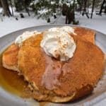 A plate of gingerbread protein pancakes made from homemade gingerbread pancake mix, with maple syrup and whipped cream, on a snow covered picnic table.