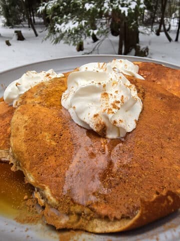 A plate of gingerbread protein pancakes made from homemade gingerbread pancake mix, with maple syrup and whipped cream, on a snow covered picnic table.