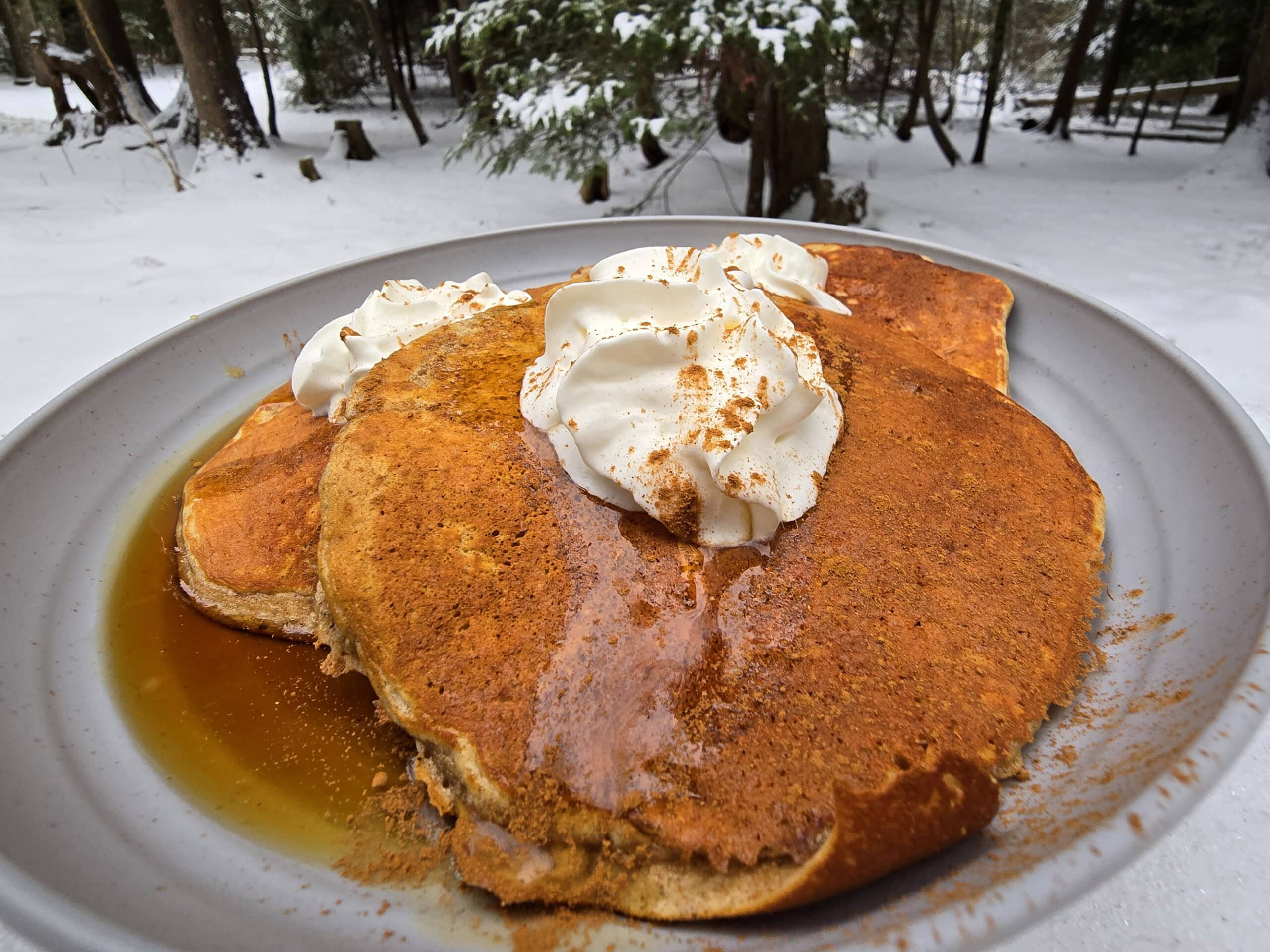 A plate of gingerbread protein pancakes made from homemade gingerbread pancake mix, with maple syrup and whipped cream,  on a snow covered picnic table.