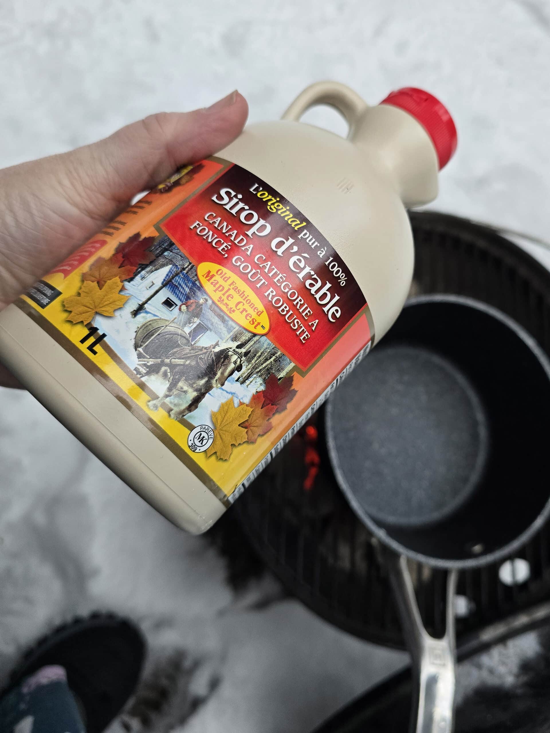 A hand holds a bottle of maple syrup over a campfire with a pot on it.