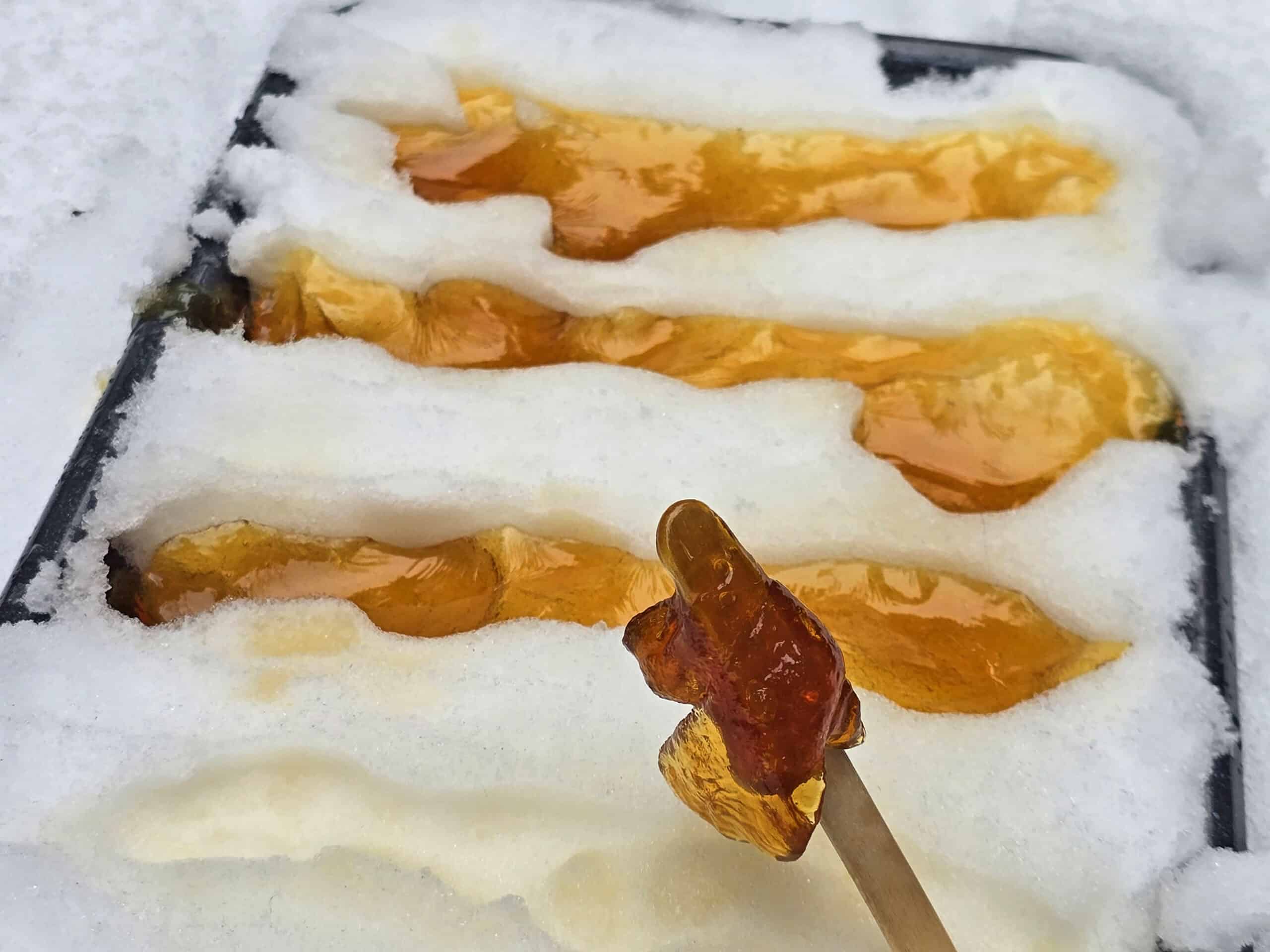 A popsicle stick with maple taffy on it, in front of a pan of maple snow taffy.