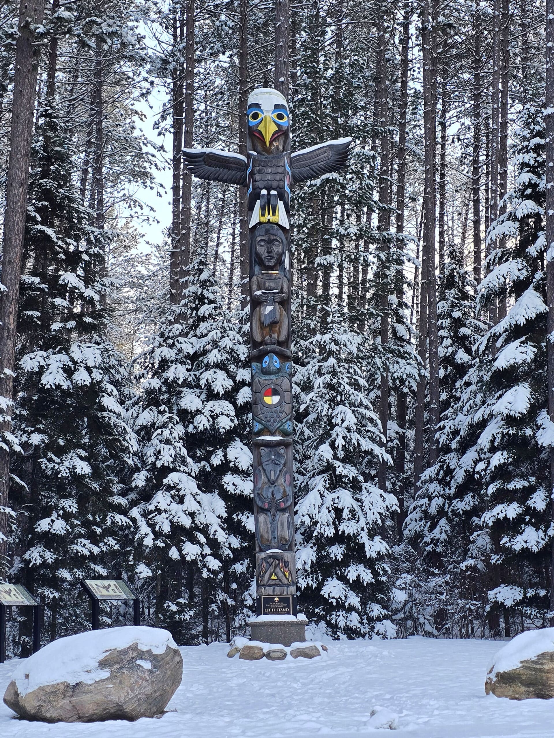 A snow covered totem pole with a winter forest behind it.