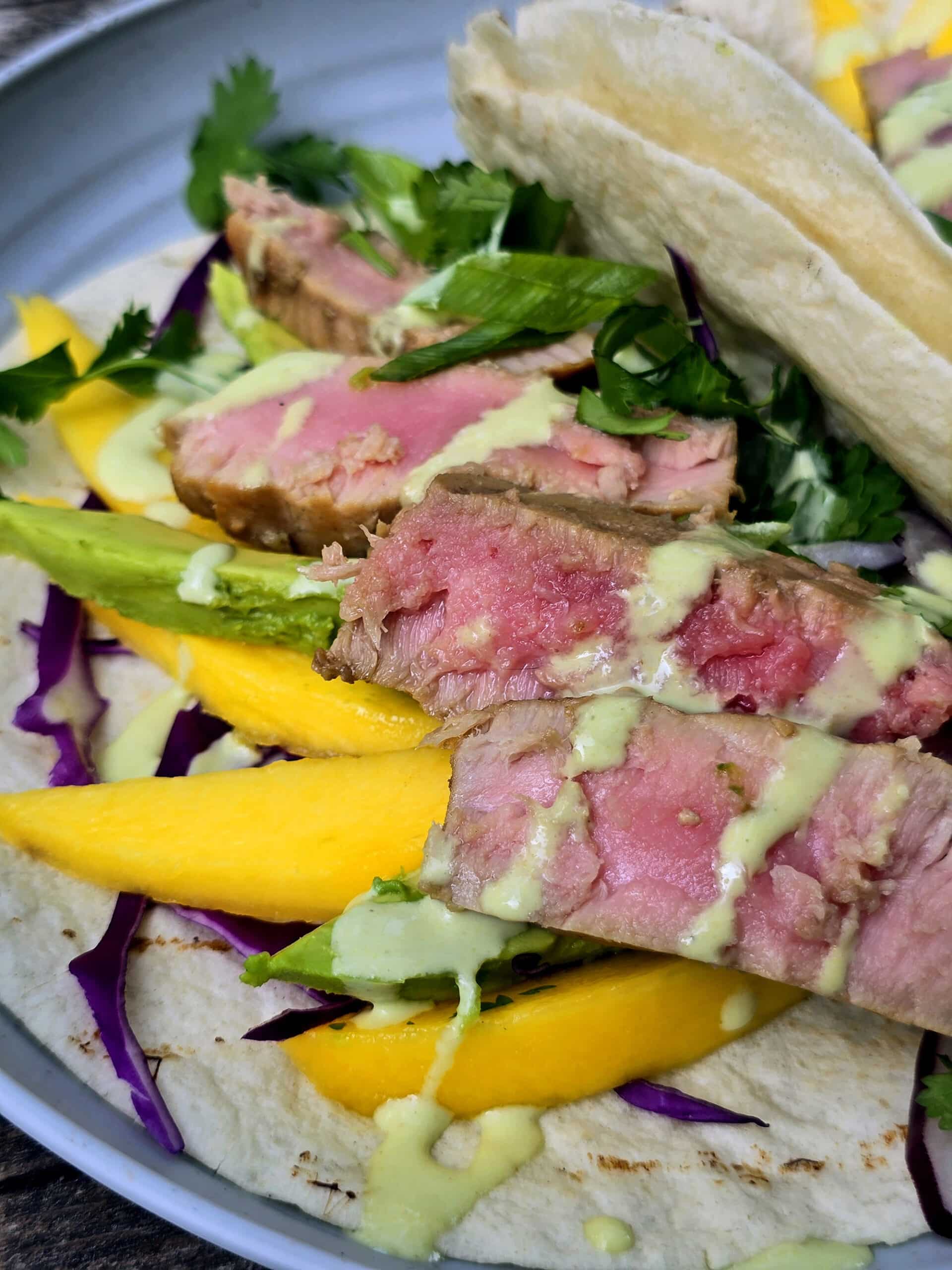 2 campfire grilled tuna tacos with mango slices, avocado, red cabbage, cilantro, and wasabi mayo on a plate.