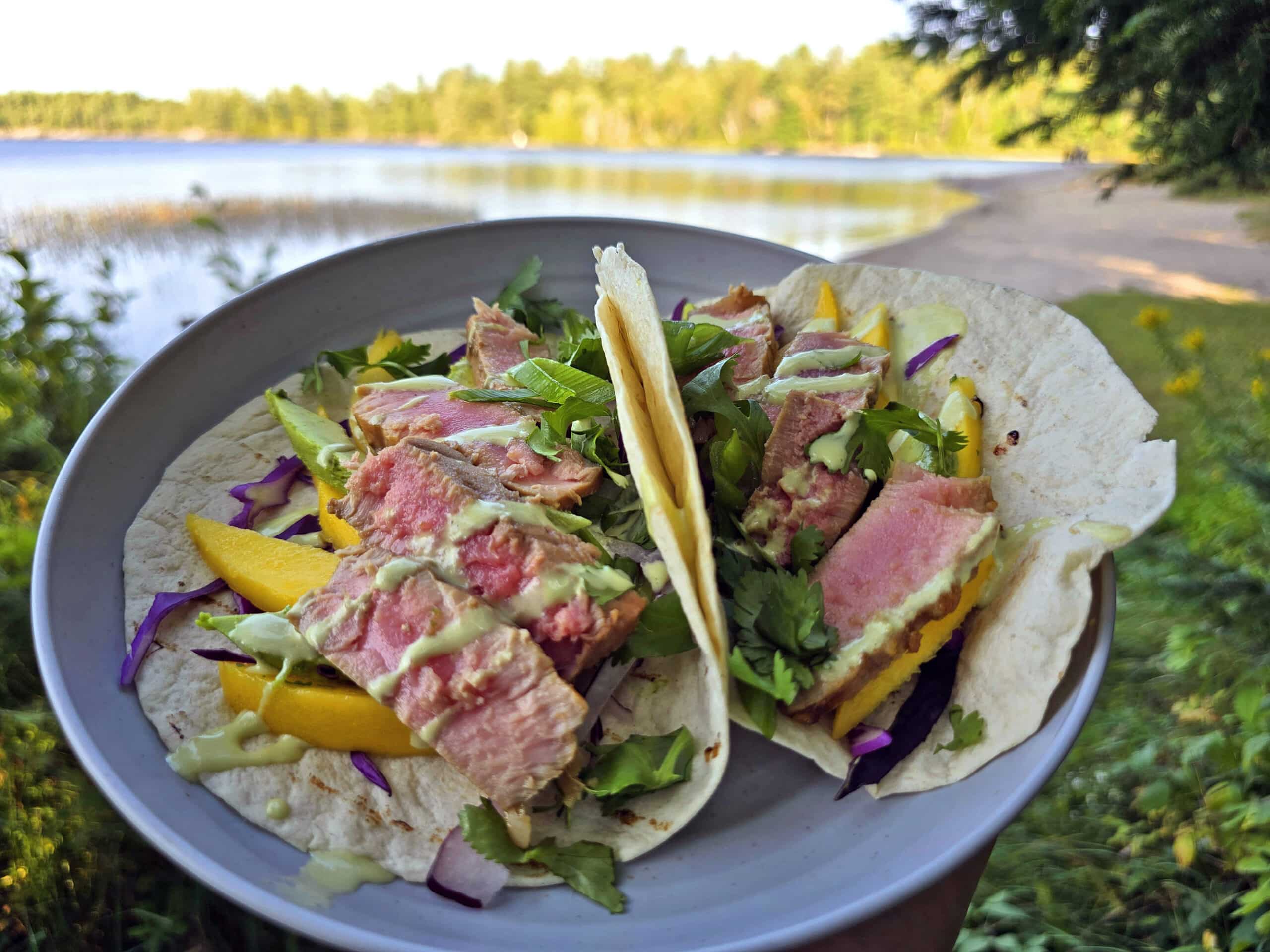 2 tacos with grilled tuna, mango slices, avocado, red cabbage, cilantro, and wasabi mayo on a plate.