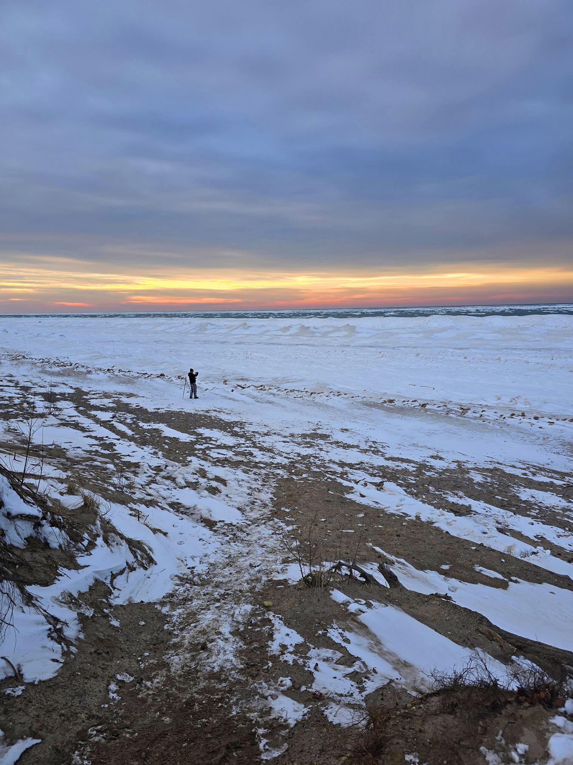 A man stands on the Pinery Provincial Park beach at sunset. It is icy and snow covered.