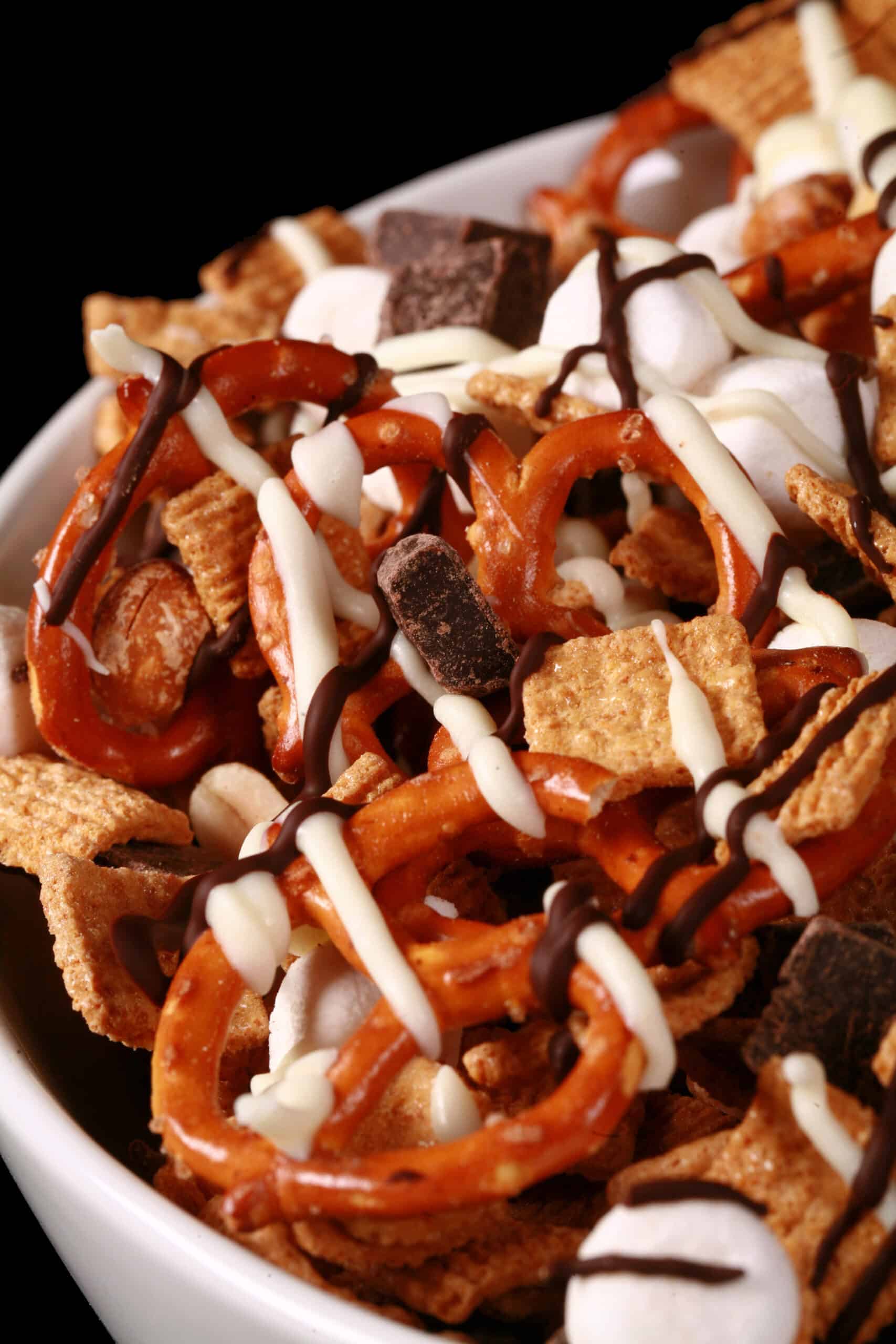 A bowl of smores trail mix, with golden grahams cereal, chocolate chunks, peanuts, pretzels, and mini marshmallows, drizzled with white and milk chocolate.