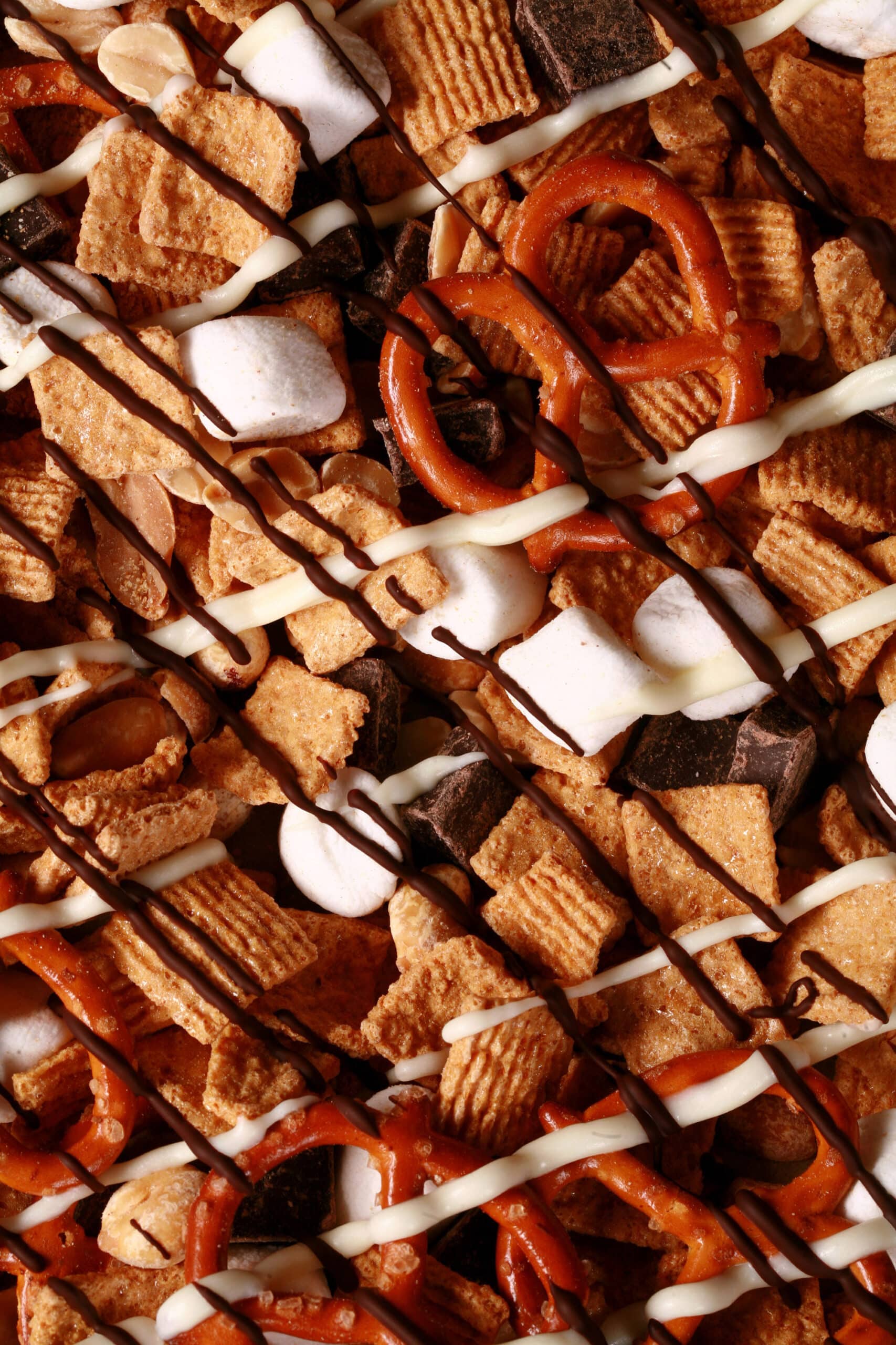 A pan of smores snack mix, with golden grahams cereal, chocolate chunks, peanuts, pretzels, and mini marshmallows, drizzled with white and milk chocolate.