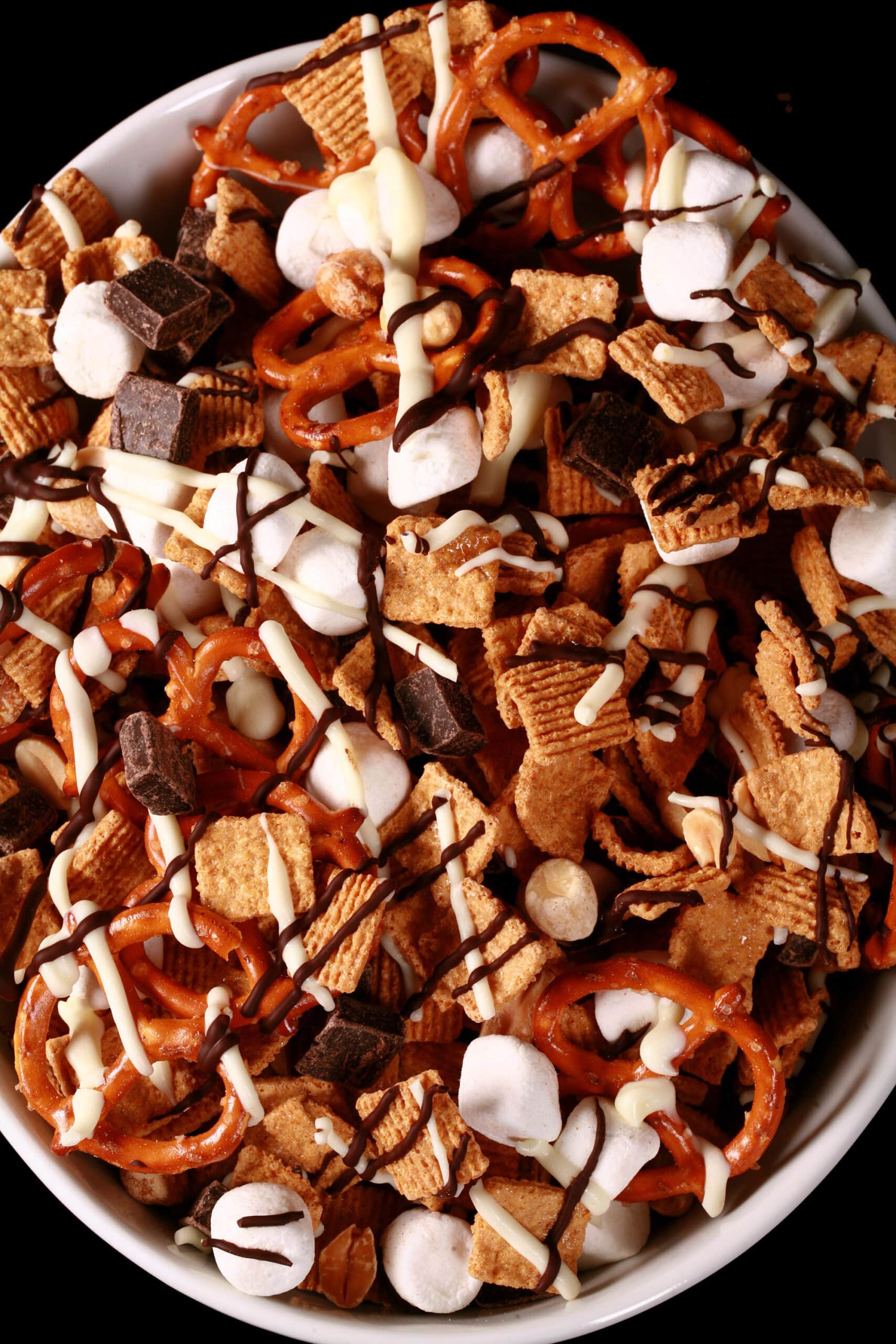 A bowl of smores trail mix, with golden grahams cereal, chocolate chunks, peanuts, pretzels, and mini marshmallows, drizzled with white and milk chocolate.