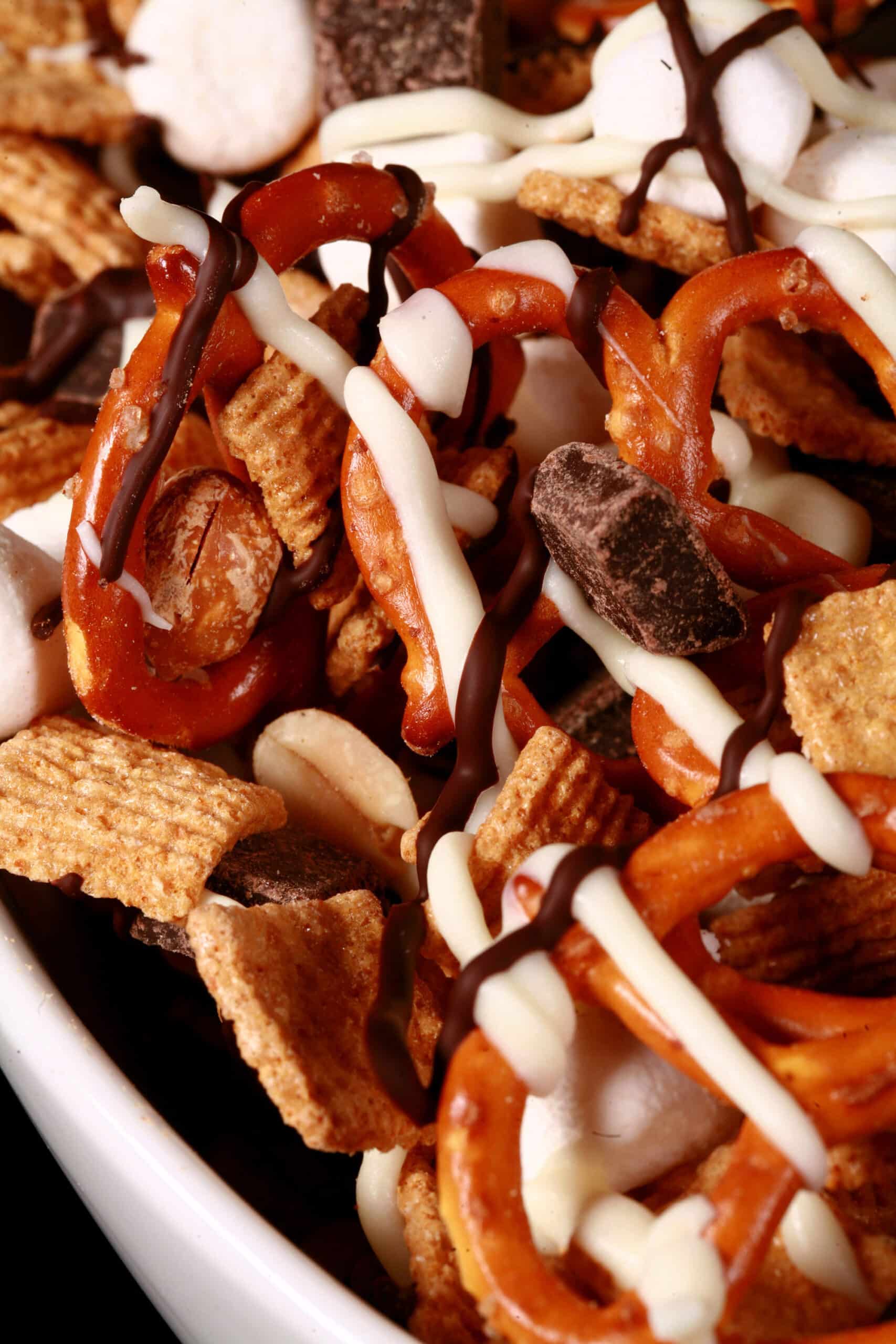 A bowl of smores trail mix, with golden grahams cereal, chocolate chunks, peanuts, pretzels, and mini marshmallows, drizzled with white and milk chocolate.