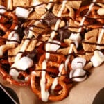 A pan of smores snack mix, with golden grahams cereal, chocolate chunks, peanuts, pretzels, and mini marshmallows, drizzled with white and milk chocolate.