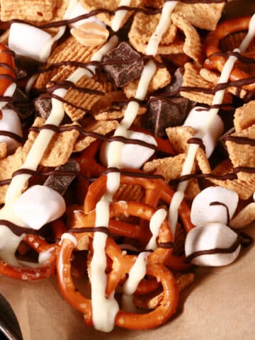 A pan of smores snack mix, with golden grahams cereal, chocolate chunks, peanuts, pretzels, and mini marshmallows, drizzled with white and milk chocolate.