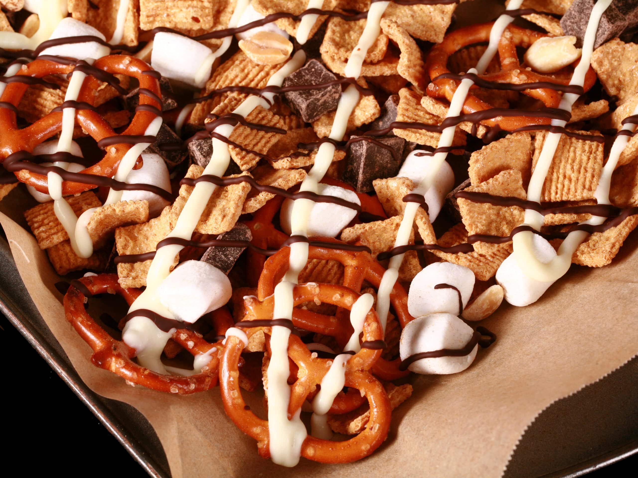 A pan of smores snack mix, with golden grahams cereal, chocolate chunks, peanuts, pretzels, and mini marshmallows, drizzled with white and milk chocolate.