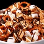 A bowl of smores trail mix, with golden grahams cereal, chocolate chunks, peanuts, pretzels, and mini marshmallows, drizzled with white and milk chocolate.