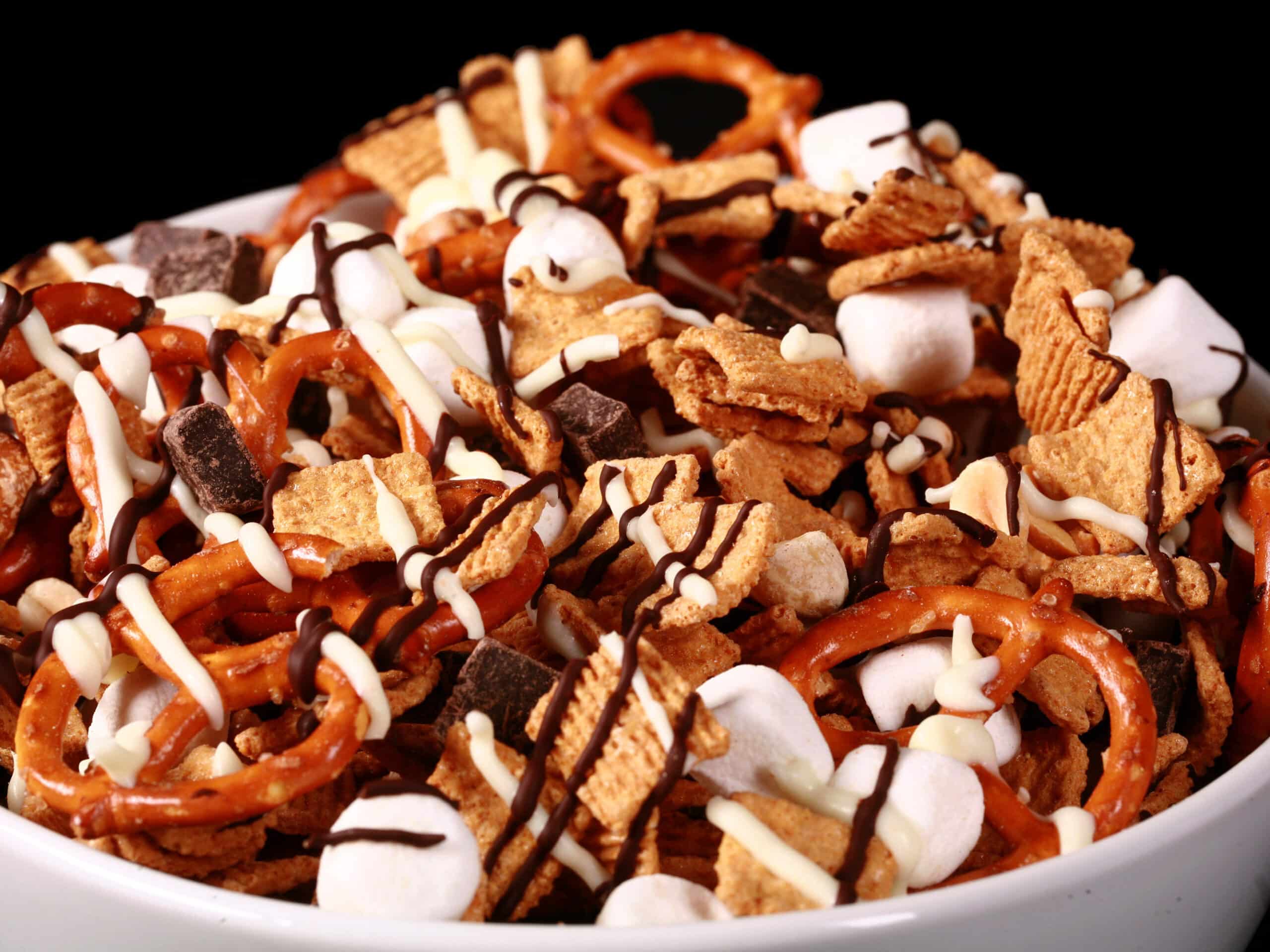 A bowl of smores trail mix, with golden grahams cereal, chocolate chunks, peanuts, pretzels, and mini marshmallows, drizzled with white and milk chocolate.