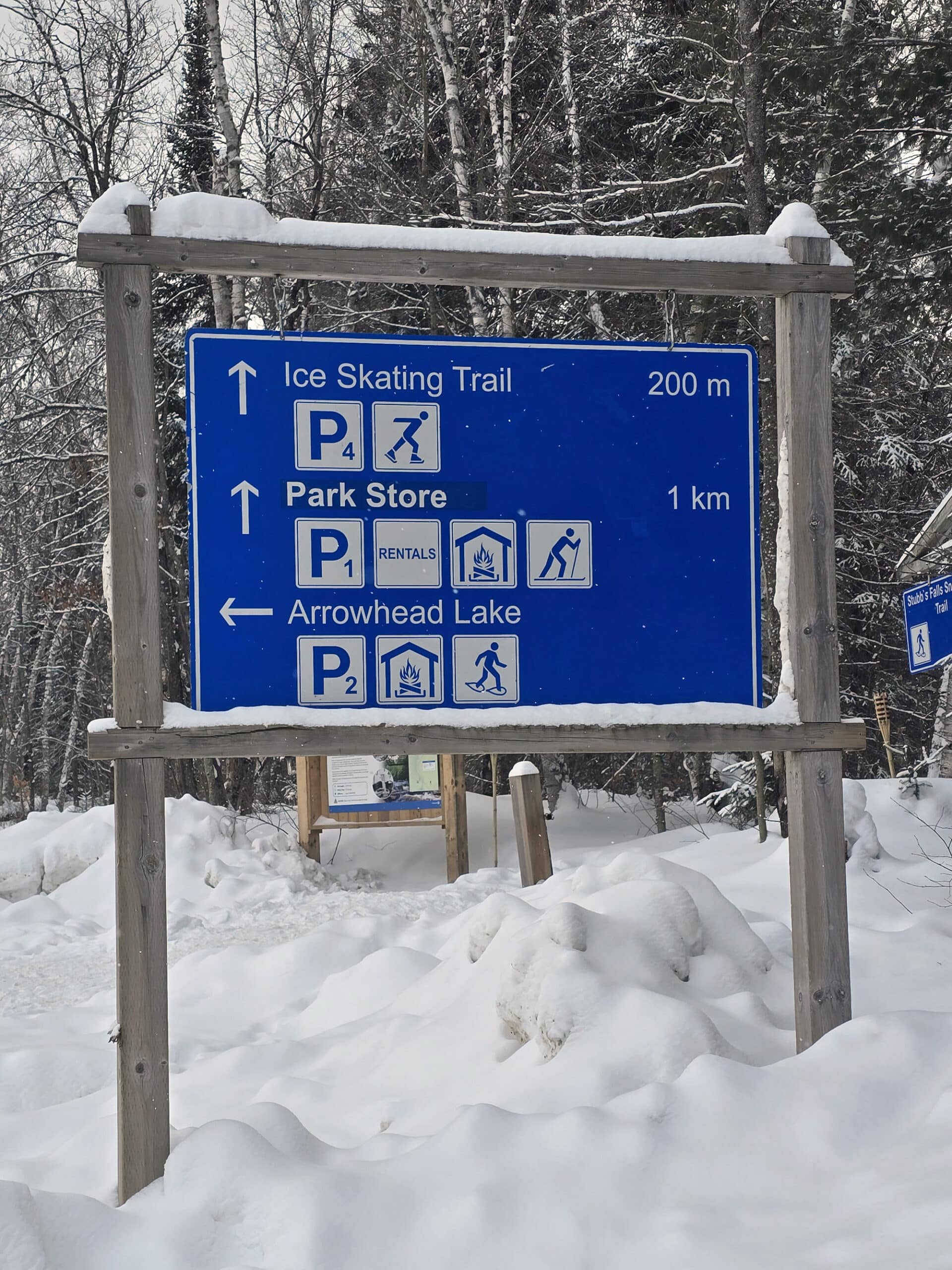 A blue and white road sign with symbols, words, and arrows directing drivers to the skating trail, park store, and arrowhead lake.