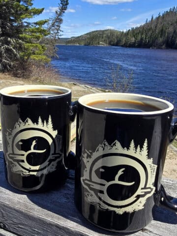 2 mugs of campfire coffee on a picnic table next to a lake.