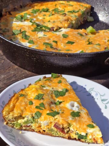 A slice of bacon, cheddar, and broccoli fritatta, in front of a pan of campfire fritatta.