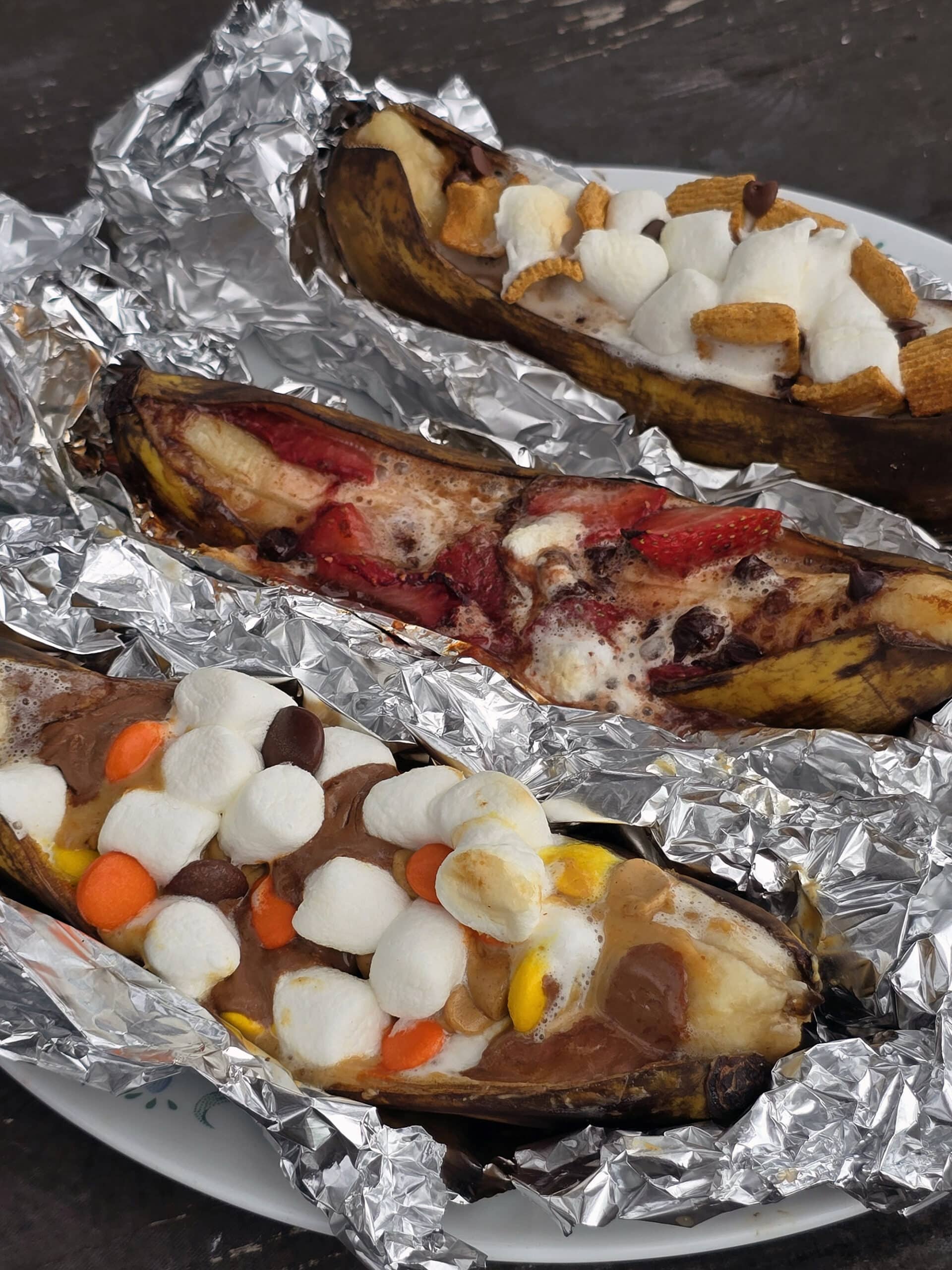 3 grilled banana boats.  One is stuffed with reeses pieces peanut butter cups and marshmallows, another with marshmallows, strawberries and chocolate chips, and the final one with marshmallows, chocolate chips, and golden grahams cereal.