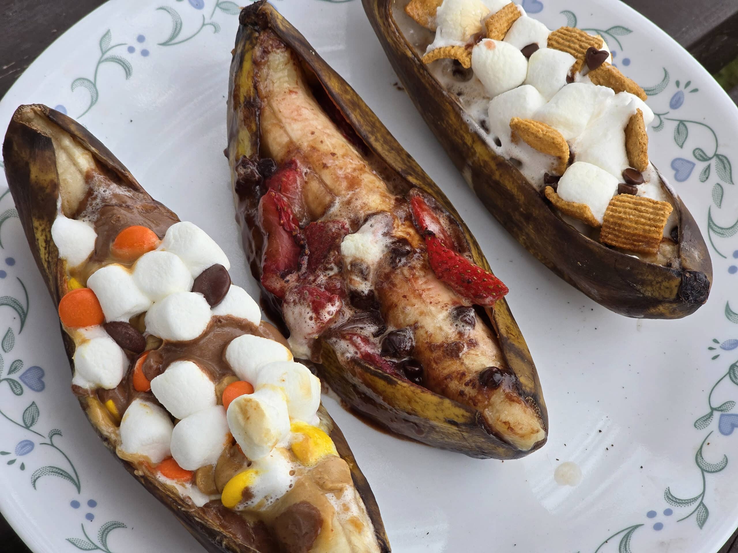 3 grilled banana boats.  One is stuffed with reeses pieces peanut butter cups and marshmallows, another with marshmallows, strawberries and chocolate chips, and the final one with marshmallows, chocolate chips, and golden grahams cereal.