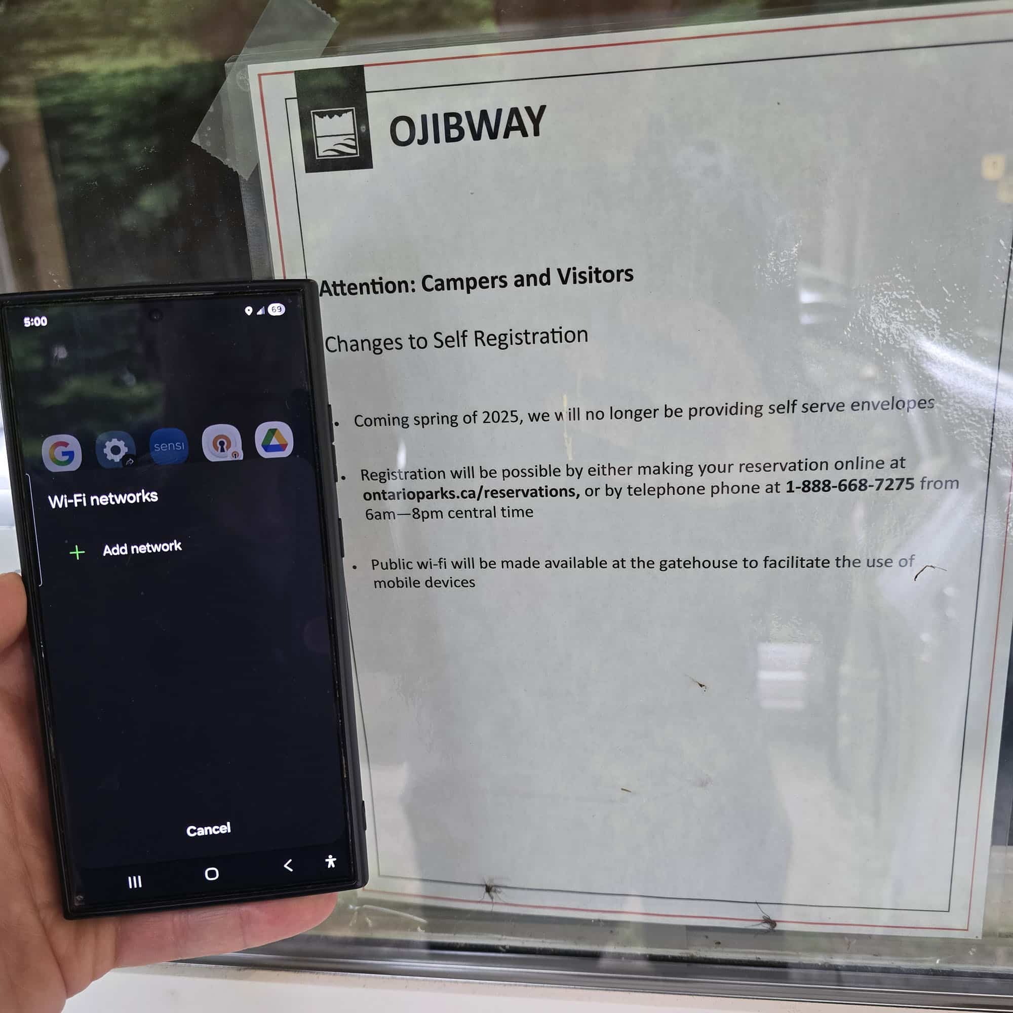 A hand holds a cell phone with no networks available next to a sign saying there's wifi.