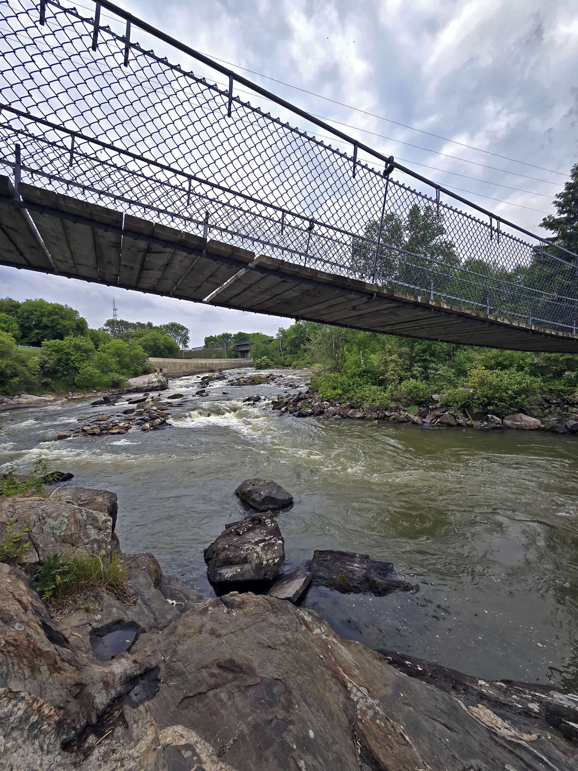 The Roy Wilson suspension bridge as seen from below and the side, with a small waterfall in the background.
